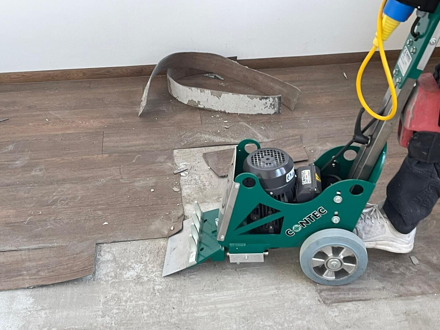 Eine Person bedient eine Maschine zum Entfernen von grünem Bodenbelag und entfernt damit braunen Vinylbodenbelag von einer Betonoberfläche. Streifen des entfernten Bodenbelags und Schutt liegen verstreut in der Umgebung.