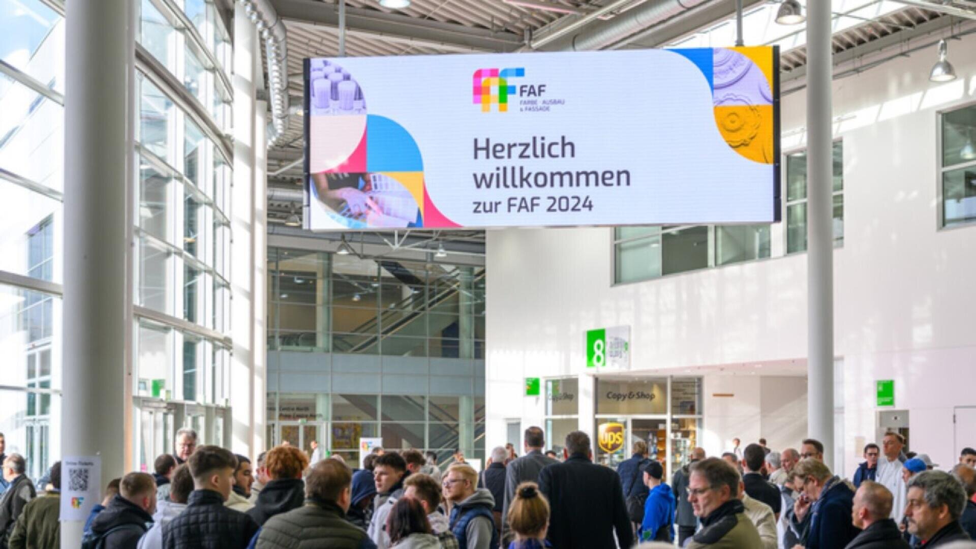 Large group of people in a modern indoor venue seen from behind, standing under a sign that reads “Herzlich willkommen zur FAF 2024” with colorful graphics, welcoming attendees to an event.