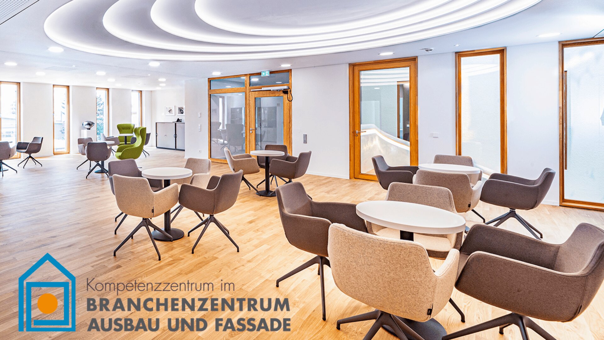 Moderne, helle Lounge mit runden Tischen und gepolsterten Stühlen auf Holzboden, großen Fenstern und minimalistischem Dekor. Der Logotext auf Deutsch lautet: Kompetenzzentrum im Branchenzentrum Ausbau und Fassade.