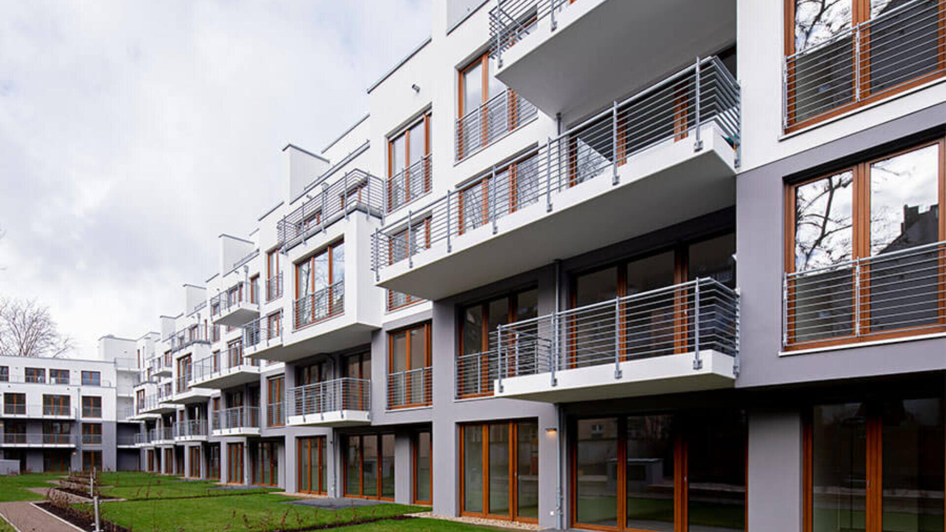 Modernes Mehrfamilienhaus mit mehreren Stockwerken, großen Glasfenstern und geräumigen Balkonen. Die Außenfassade ist weiß mit grauen Akzenten. Vor dem Gebäude befinden sich eine Rasenfläche und ein Gehweg.