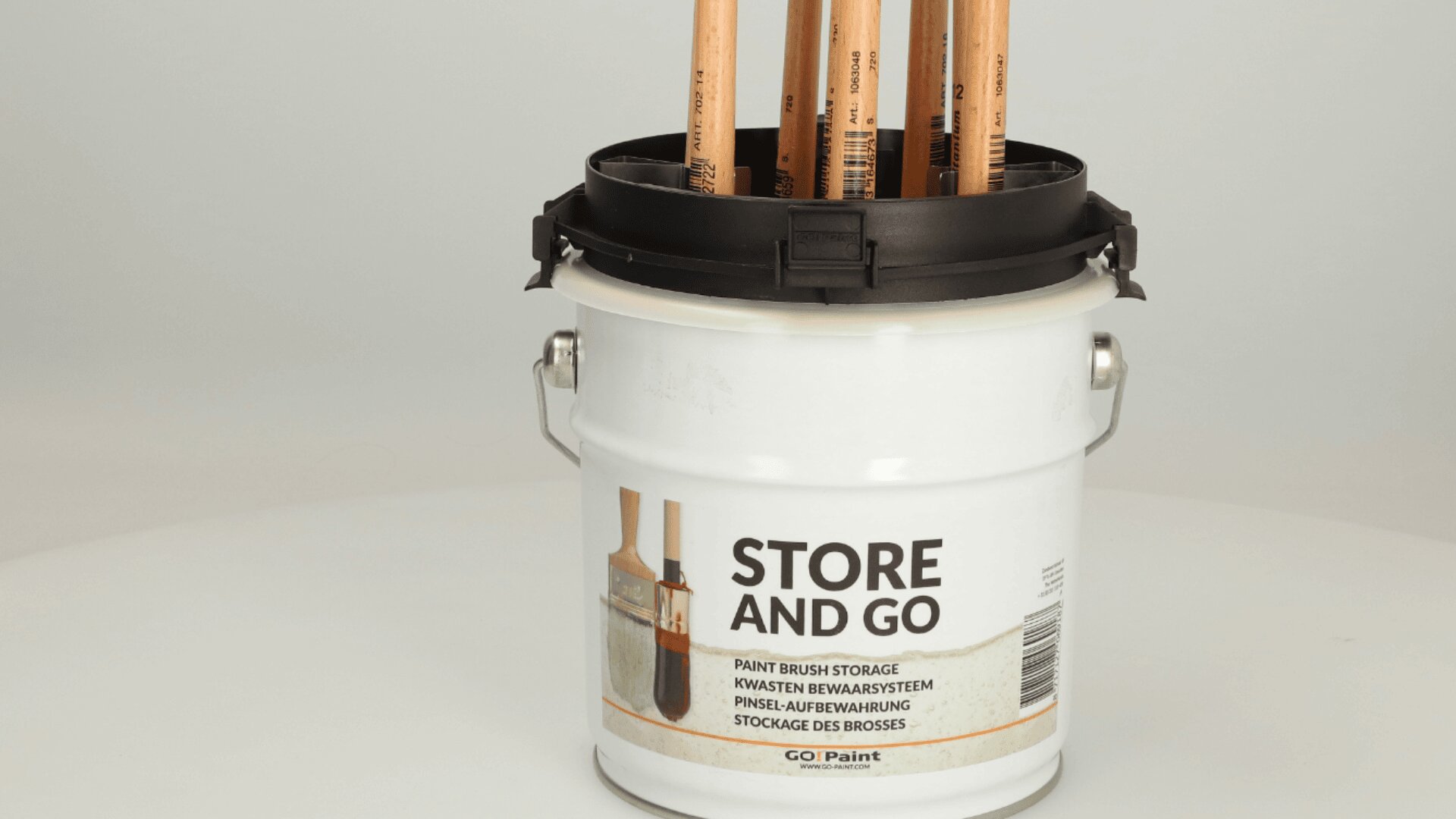 A white paint bucket labeled STORE AND GO holds several paintbrushes upright inside a black storage lid designed for organizing and storing brushes.