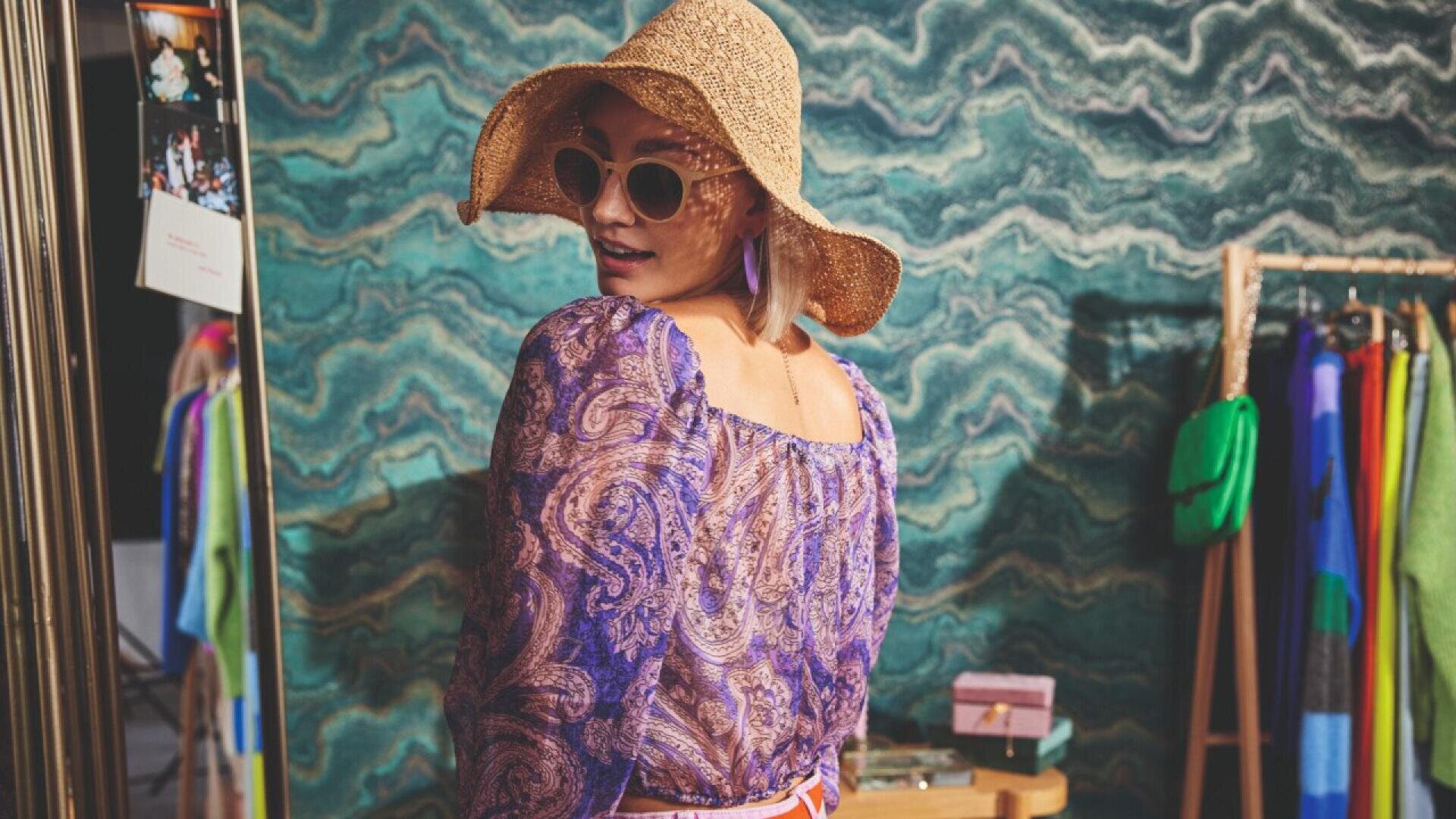 A woman wearing a wide-brimmed straw hat, sunglasses, and a purple paisley top stands indoors, looking over her shoulder. Colorful clothing and a wavy green patterned wall are visible in the background.