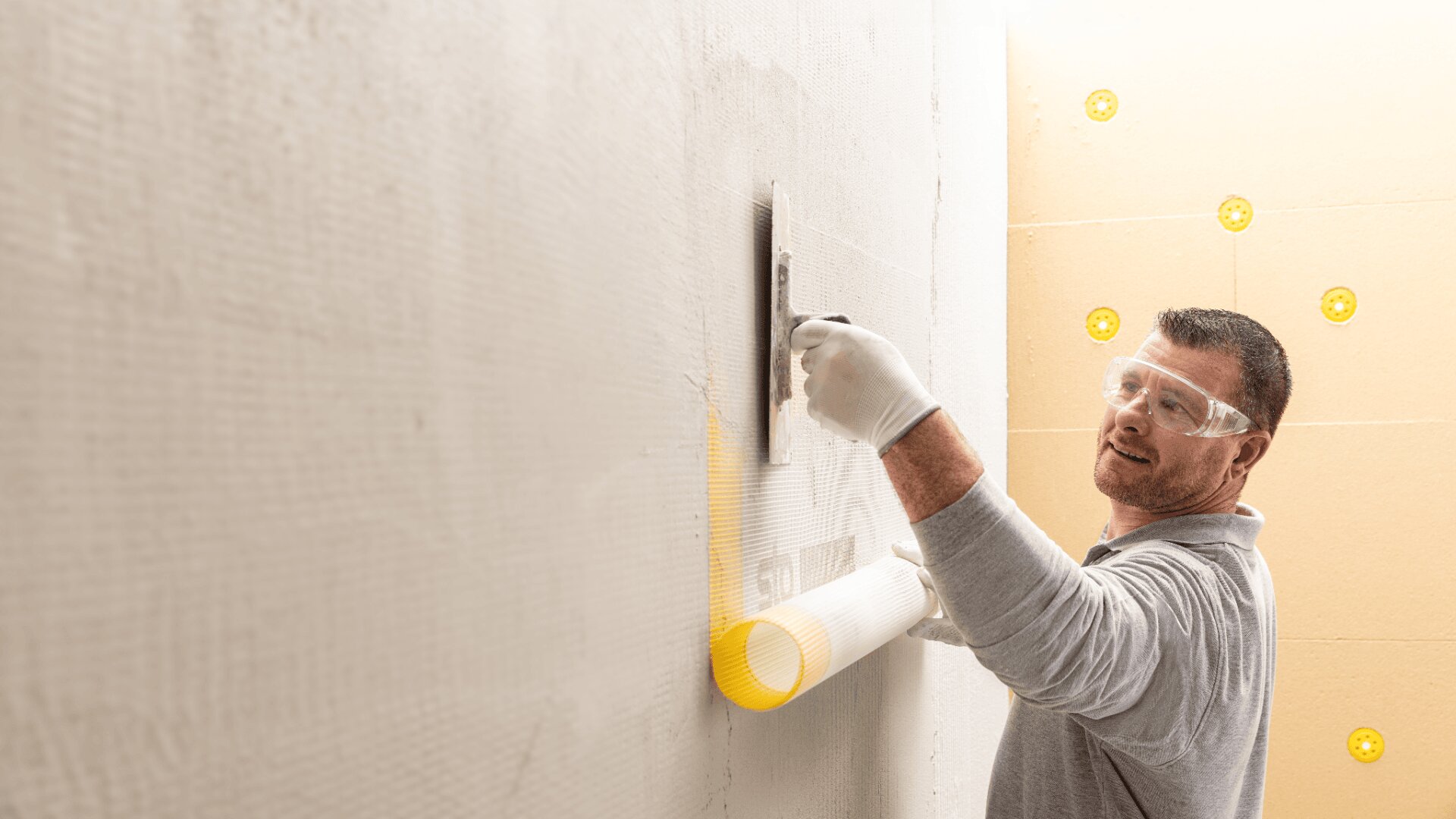 Ein Mann mit Schutzbrille und Handschuhen glättet mit einer Kelle Putz oder Kleber auf einer Wand. In der Nähe befindet sich eine Rolle Netzgewebe. An der angrenzenden Wand sind gelbe Befestigungselemente zu sehen.