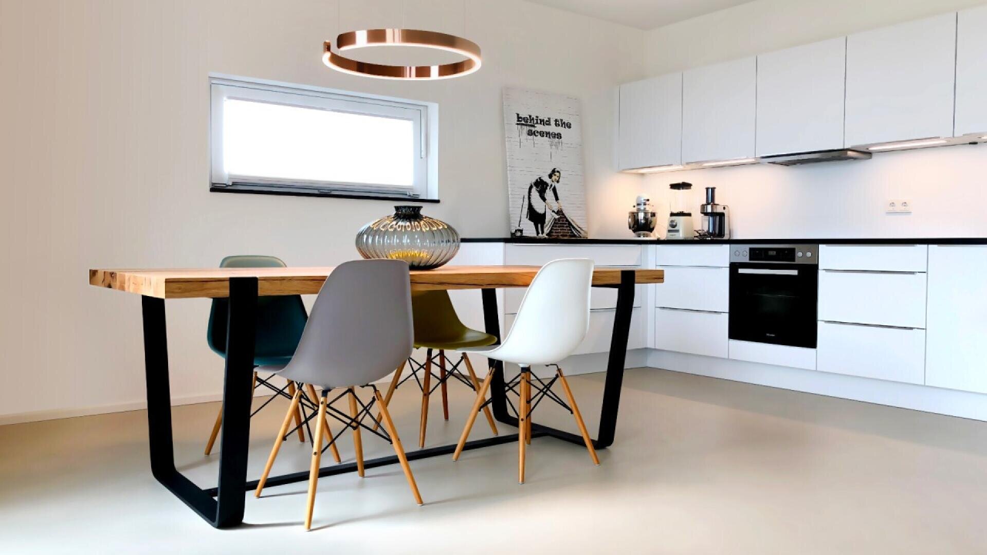A modern kitchen with white cabinets, a wooden dining table, four colorful chairs, a large glass vase, a circular pendant light, and wall art above the countertop.