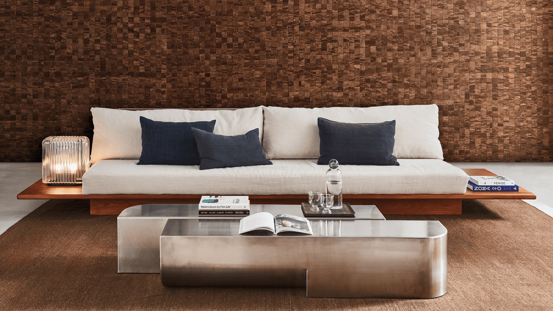 Ein modernes Wohnzimmer verfügt über ein niedriges Holzsofa mit weißen und dunkelblauen Kissen, einen Couchtisch aus Metall mit Büchern und Glaswaren, eine braun strukturierte Wand und eine Lampe mit einem Schirm aus geripptem Glas.