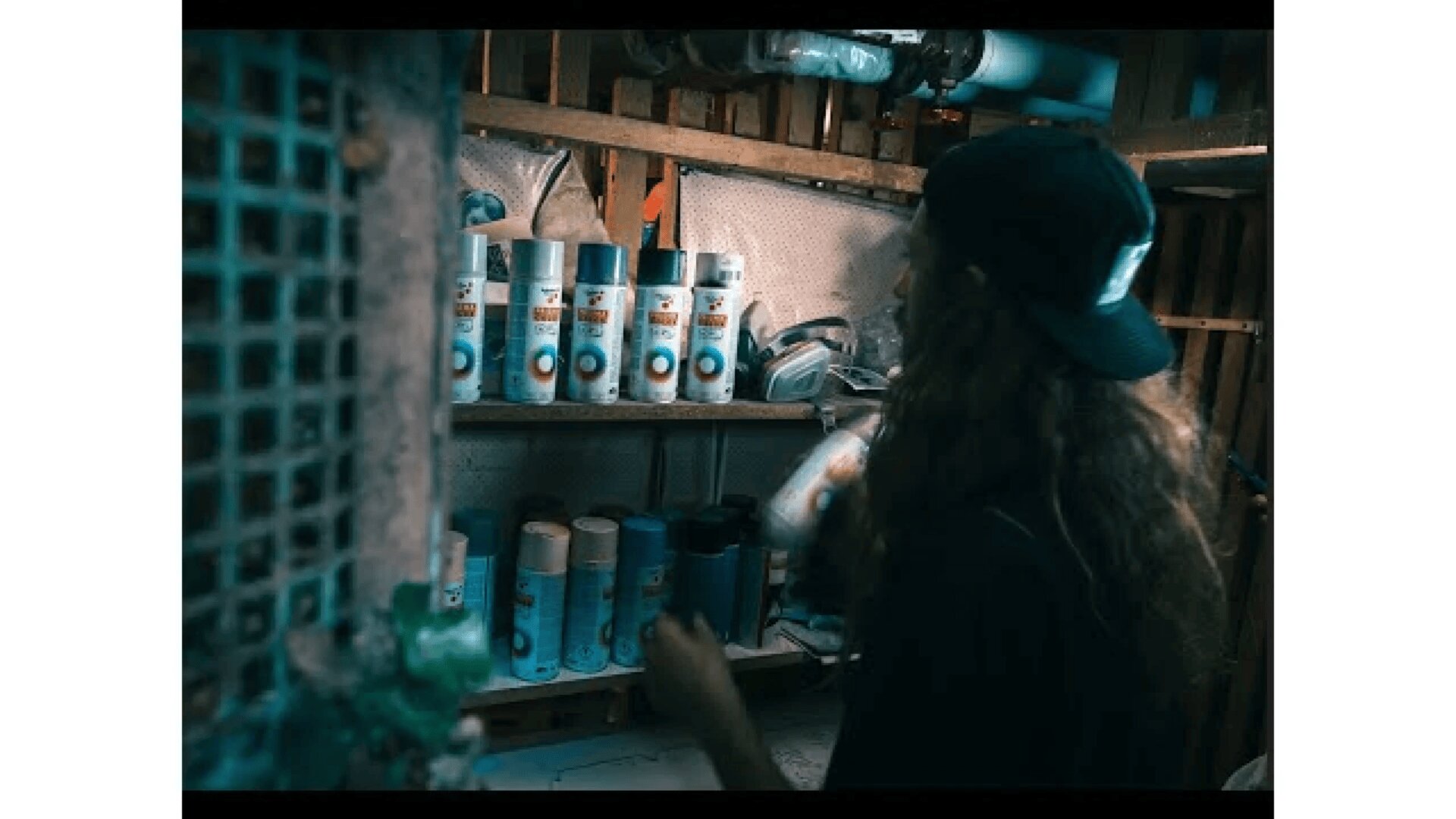 A person with long hair and a backwards cap stands in a dimly lit room, holding a spray paint can and looking at shelves stocked with various spray paint cans and protective gear.