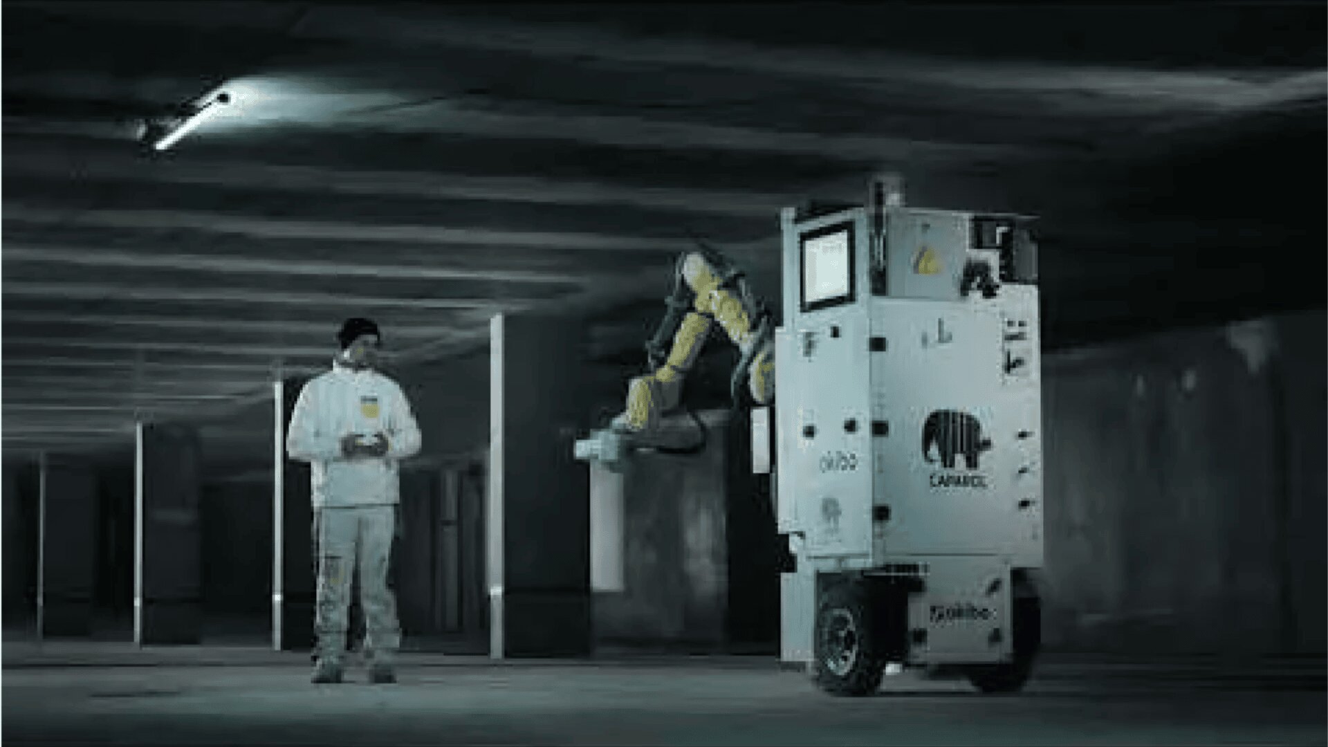 Eine Person in weißer Arbeitskleidung steht in einem schwach beleuchteten Parkhaus und bedient eine große Robotermaschine mit der Aufschrift „Canvas“, die Farbe auf eine Betondecke sprüht.