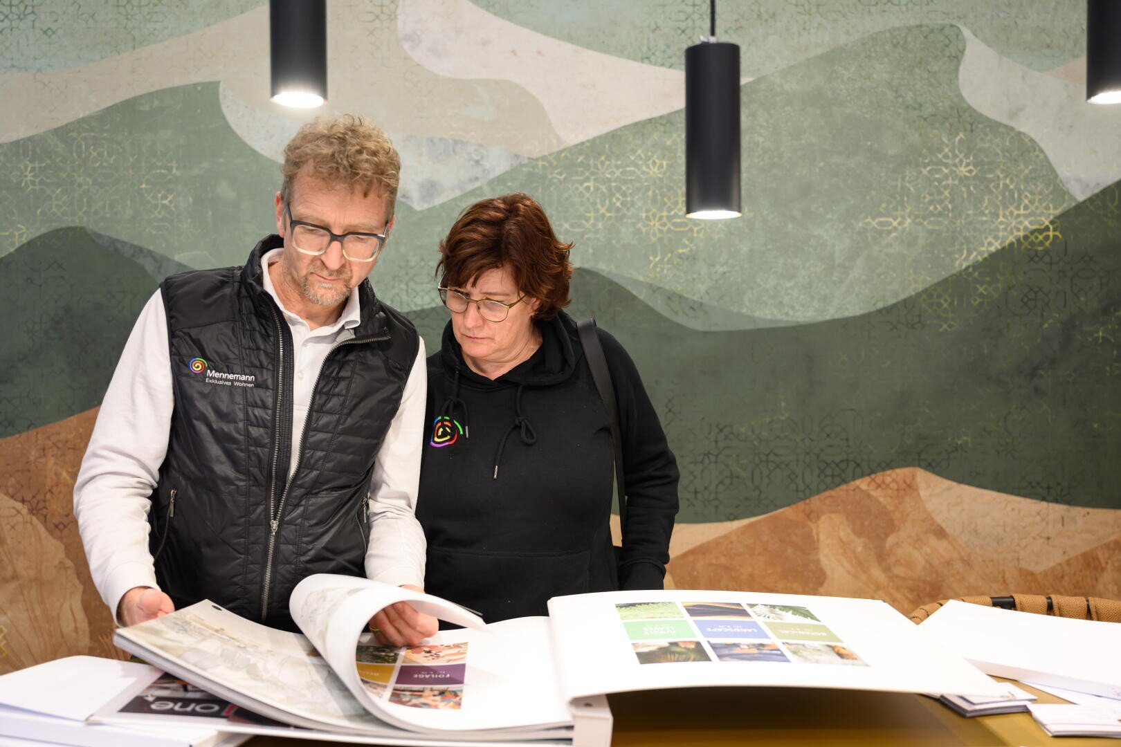 Zwei Personen stehen an einem Schreibtisch und blättern in großen Büchern oder Katalogen mit gedruckten Seiten. Sie wirken konzentriert und tragen legere Arbeitskleidung. Hinter ihnen liegt ein gemusterter grün-brauner Hintergrund.