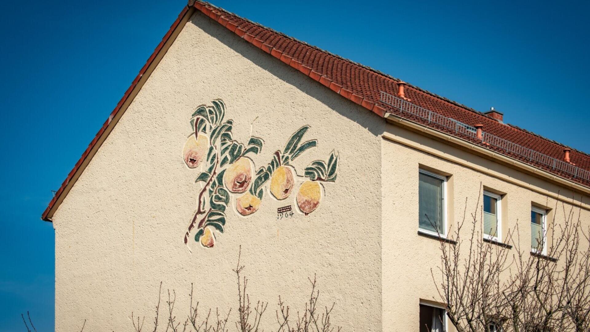 A beige house with a red-tiled roof features a mural of a branch with yellow peaches and green leaves painted on its side wall under a clear blue sky.