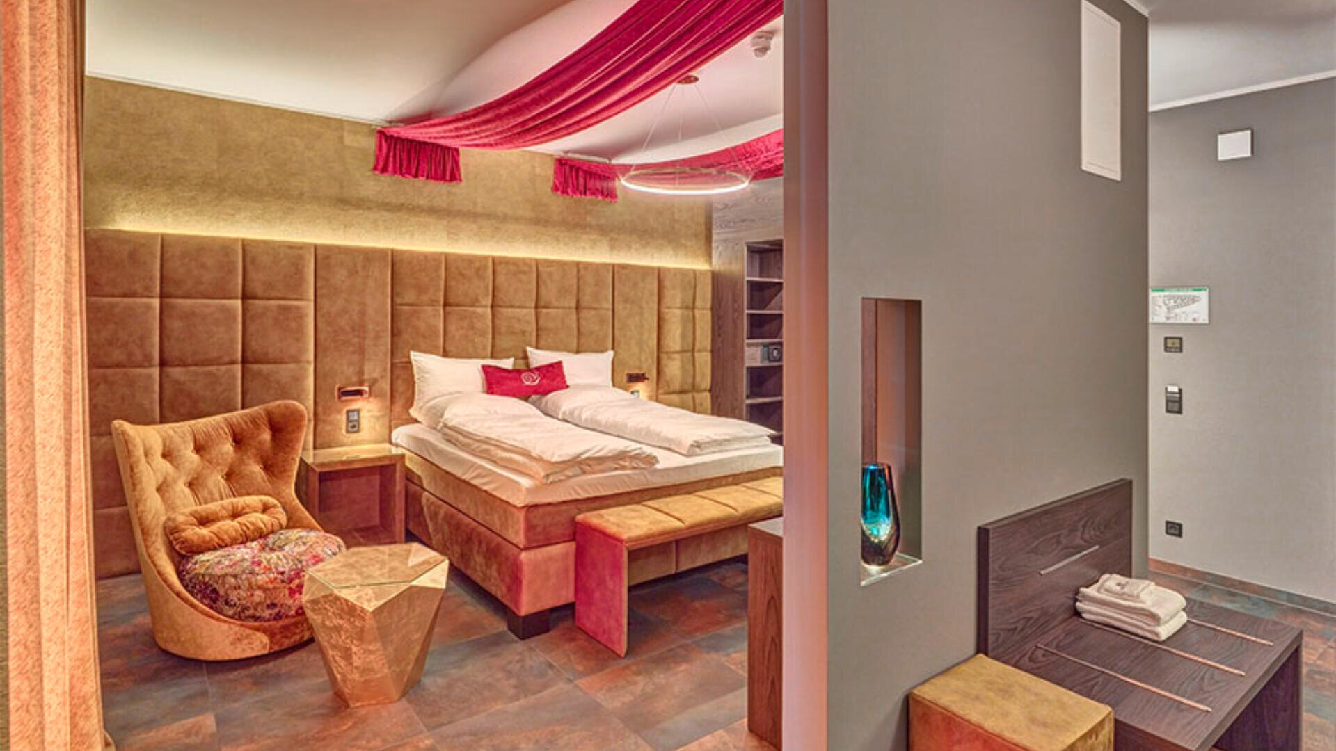 Ein stilvolles Hotelzimmer mit einem großen Bett, einem Kopfteil aus Samt, einem goldenen Sessel, einem geometrischen Beistelltisch, moderner Einrichtung und einem roten Stoffdrap an der Decke; Handtücher und eine blaue Vase stehen auf einem Regal im Vordergrund.
