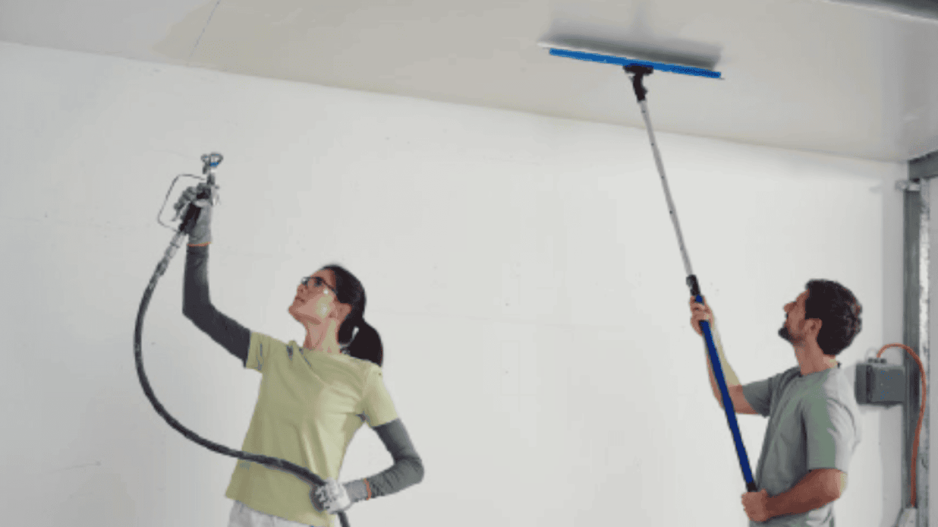 Two people working indoors; one uses a spray gun to paint or apply texture to the ceiling, while the other smooths the ceiling with a long-handled tool. Both wear casual clothing and safety glasses.