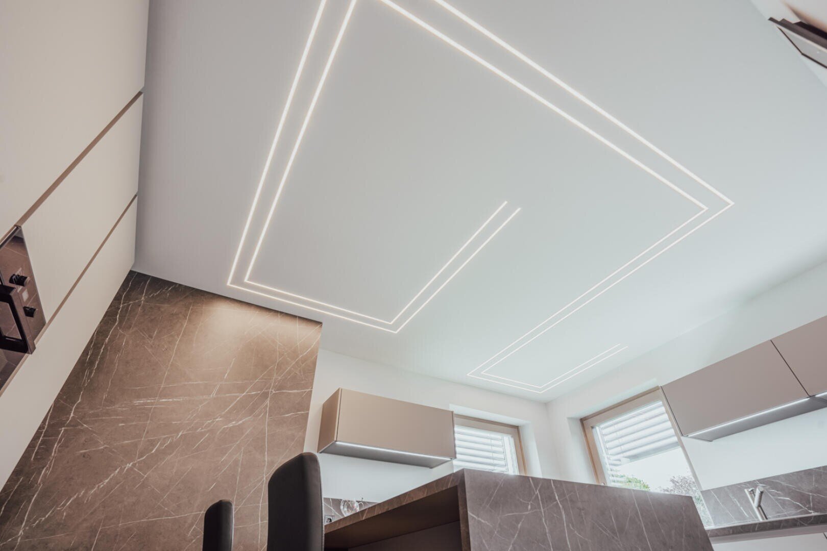 Modernes Kücheninterieur mit Marmorwand, eleganten Schränken und einer Decke mit geometrischen LED-Lichtleisten. Das Foto wurde aus einem niedrigen Winkel aufgenommen, wodurch die modernen Designelemente hervorgehoben werden.