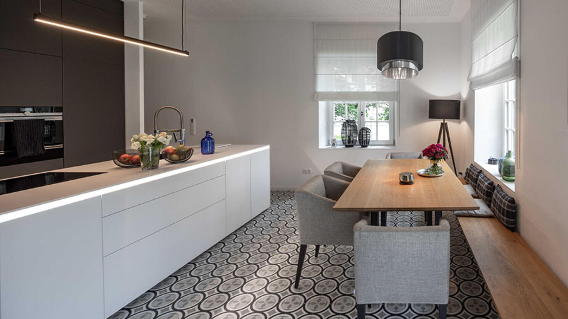 Moderne Küche und Essbereich mit gemustertem Fliesenboden, eleganten weißen Schränken, einem eingebauten Ofen, Pendelleuchten, einem hölzernen Esstisch mit grauen Stühlen und einer Fensterbank mit Kissen.