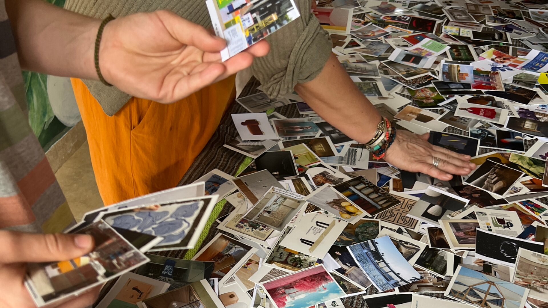 Two people sort through a large collection of small, colorful photographs spread out across a table, each holding and examining some of the photos. Only their hands and arms are visible.