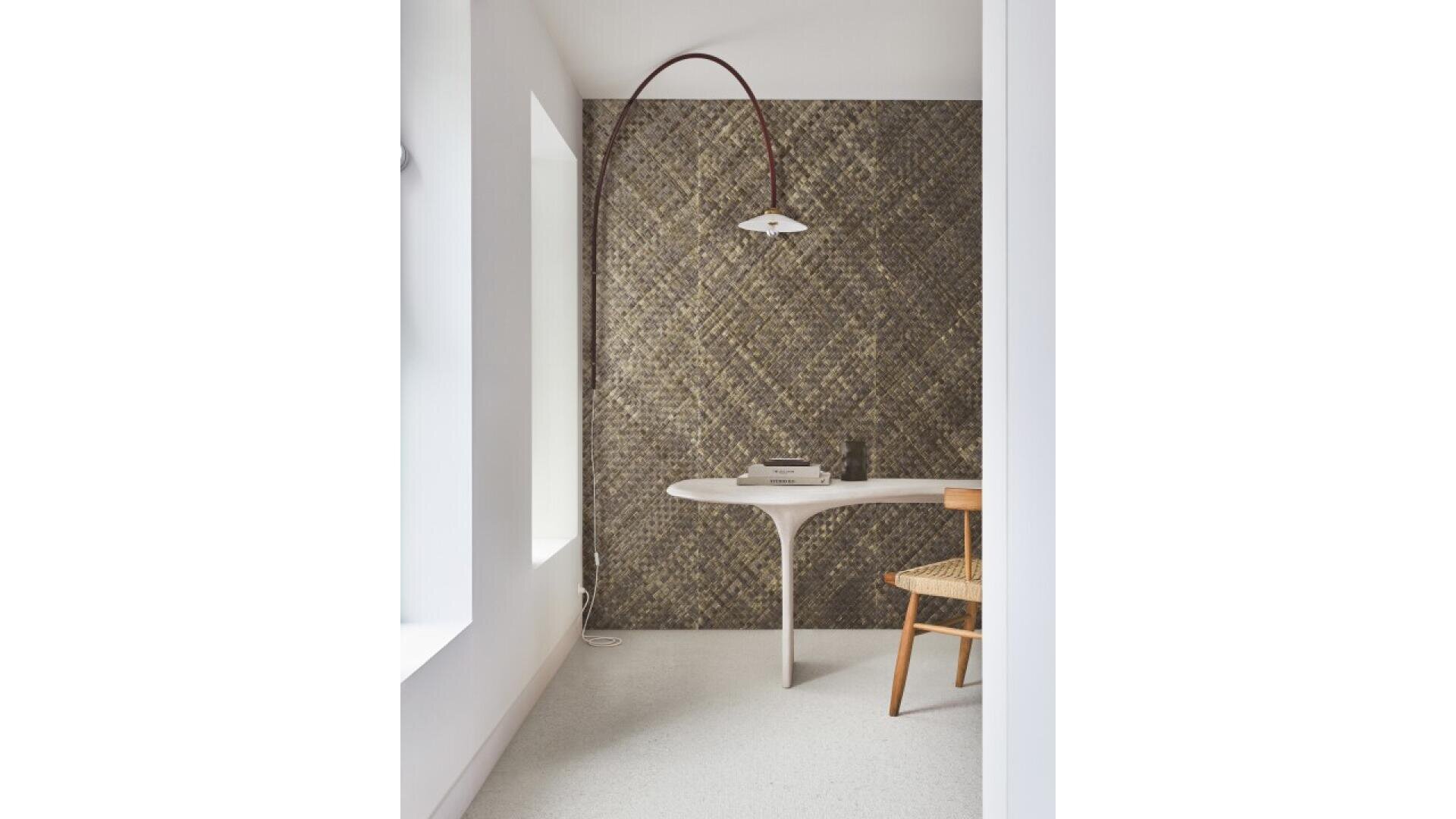 A minimalist room with a curved white desk, a woven chair, a textured gold accent wall, and a modern arc floor lamp. Natural light enters through large windows on the left.