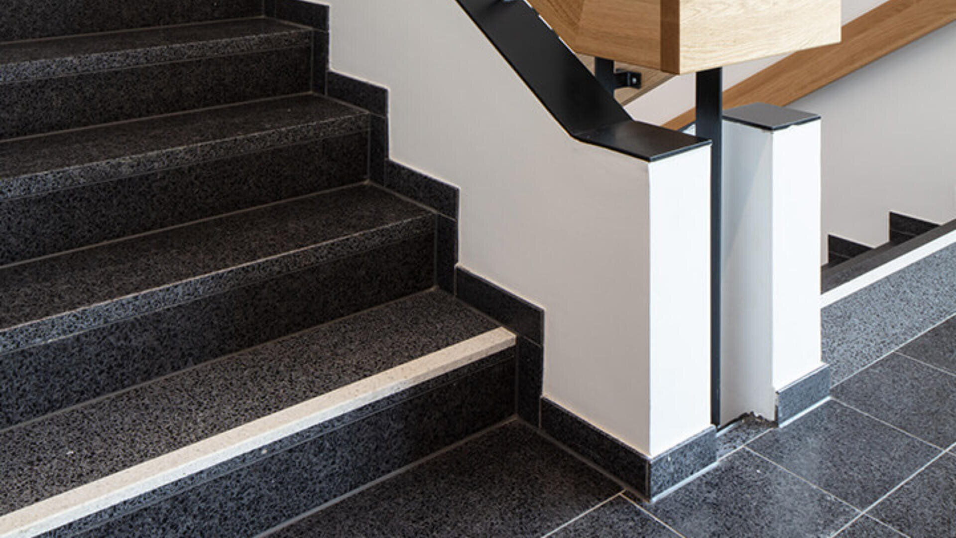 Eine moderne Innentreppe mit dunklen, polierten Steinstufen, einer weißen Wand und einem Holzhandlauf mit schwarzen Metallhalterungen. Die Treppe hat einen hellen Streifen am Rand für bessere Sichtbarkeit.
