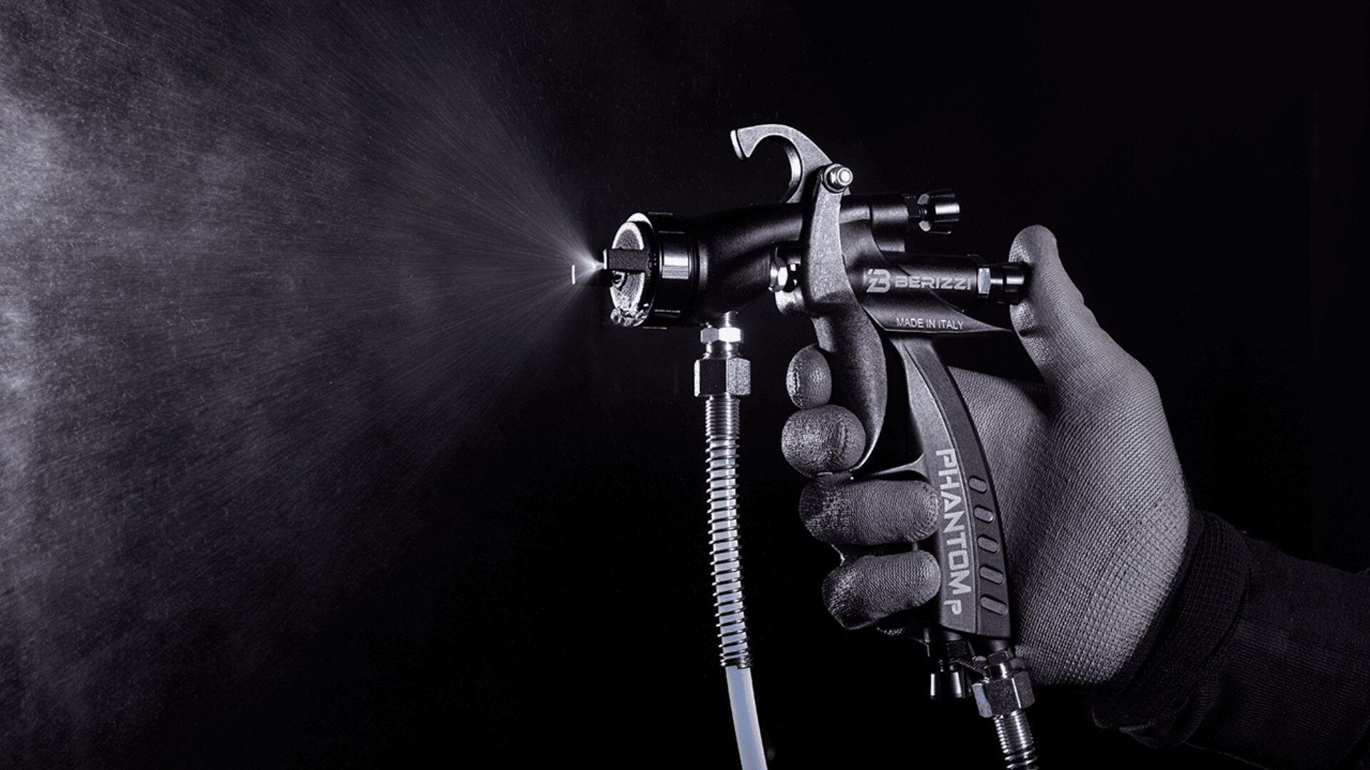 A gloved hand holds and operates a paint spray gun, releasing a fine mist of paint against a dark background.