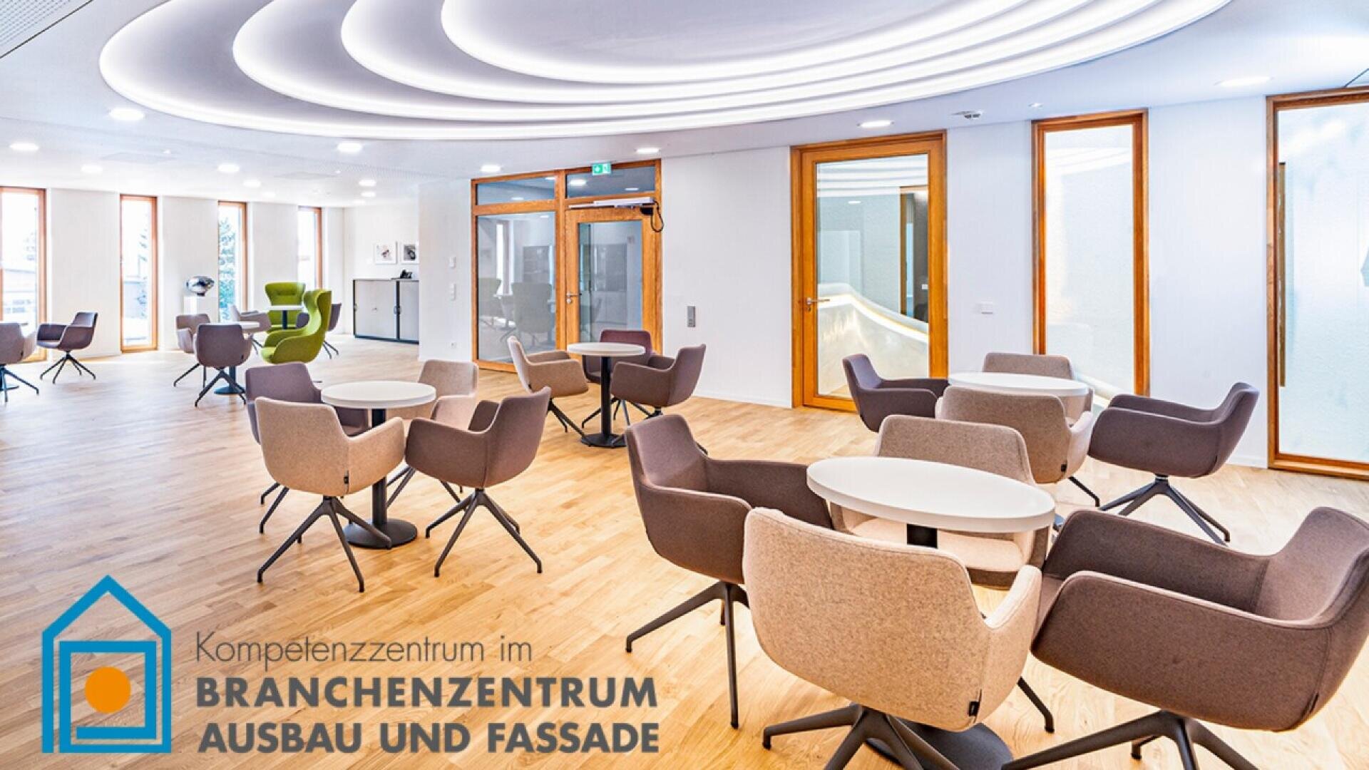 A modern, spacious lounge area with round tables and various gray and beige chairs on a light wood floor. Large windows line the walls, and the ceiling has circular recessed lighting. Logo: Branchenzentrum Ausbau und Fassade.