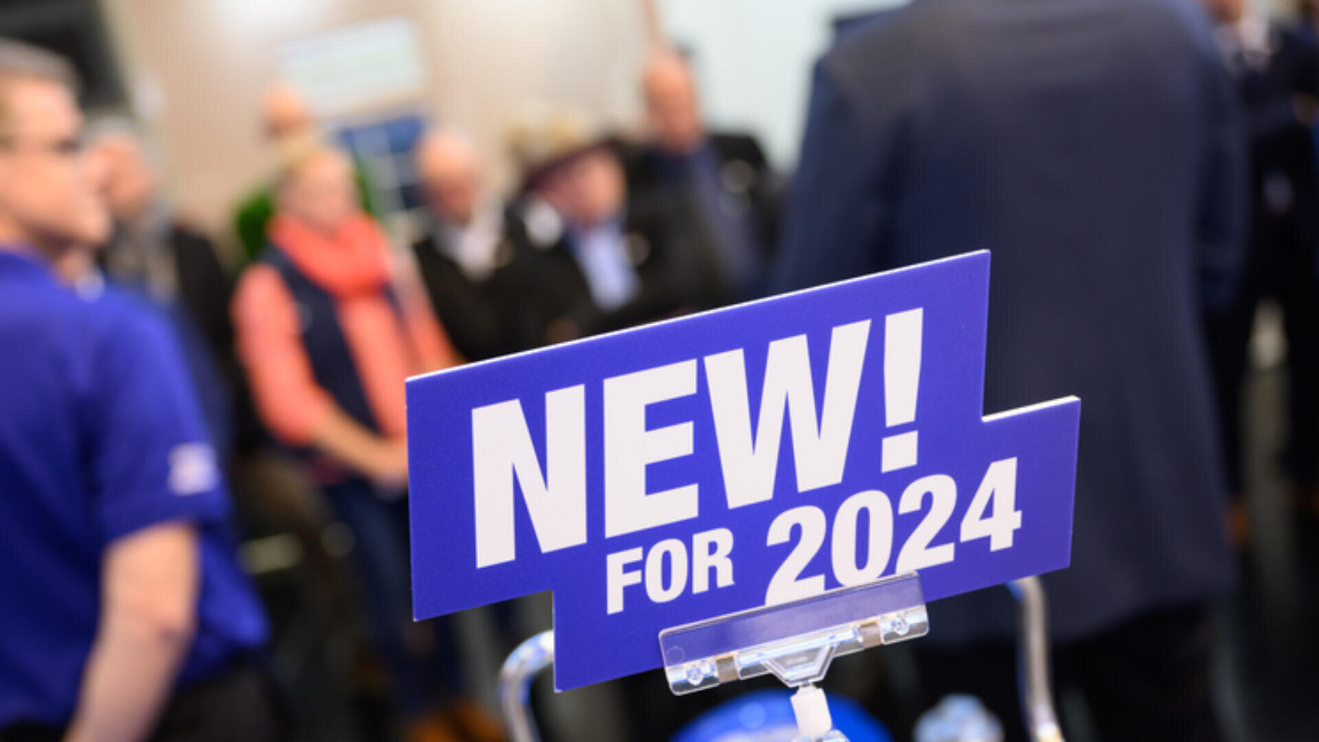 Im Vordergrund ist deutlich ein blaues Schild mit der Aufschrift „NEU! FÜR 2024“ zu sehen, im Hintergrund ist eine Gruppe von Menschen unscharf zu erkennen, was auf eine öffentliche Veranstaltung oder Ausstellung schließen lässt.