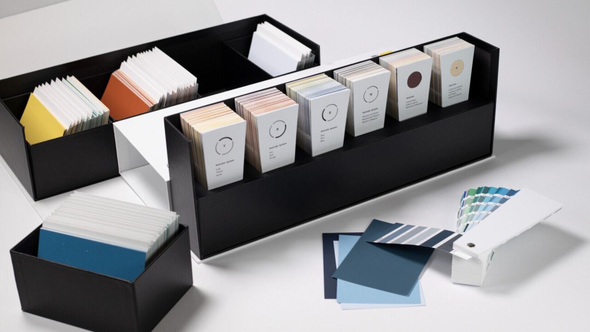 Boxes with neatly organized color swatches and paint sample cards are displayed on a white surface, along with a color fan deck and several blue color cards spread out.