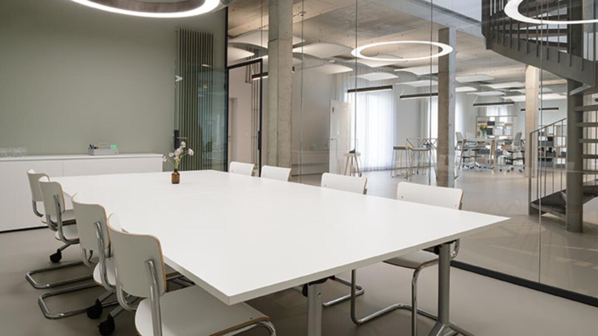 A modern conference room with a white rectangular table surrounded by white chairs, minimalist decor, glass walls, circular ceiling lights, and a view of an open office space with a spiral staircase.
