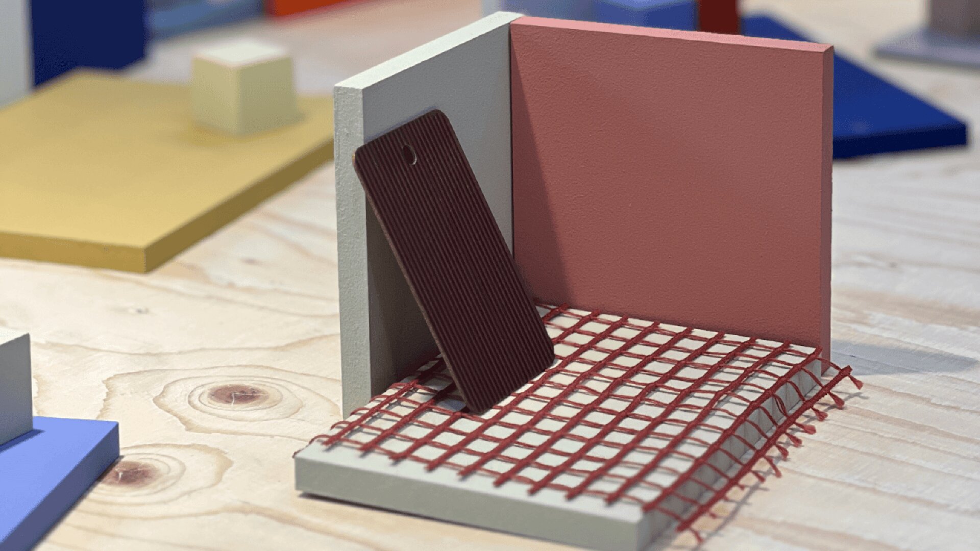 A brown rectangular tag leans against a pink and gray corner backdrop, with a red grid mesh on a wooden surface. Colorful geometric shapes are blurred in the background.