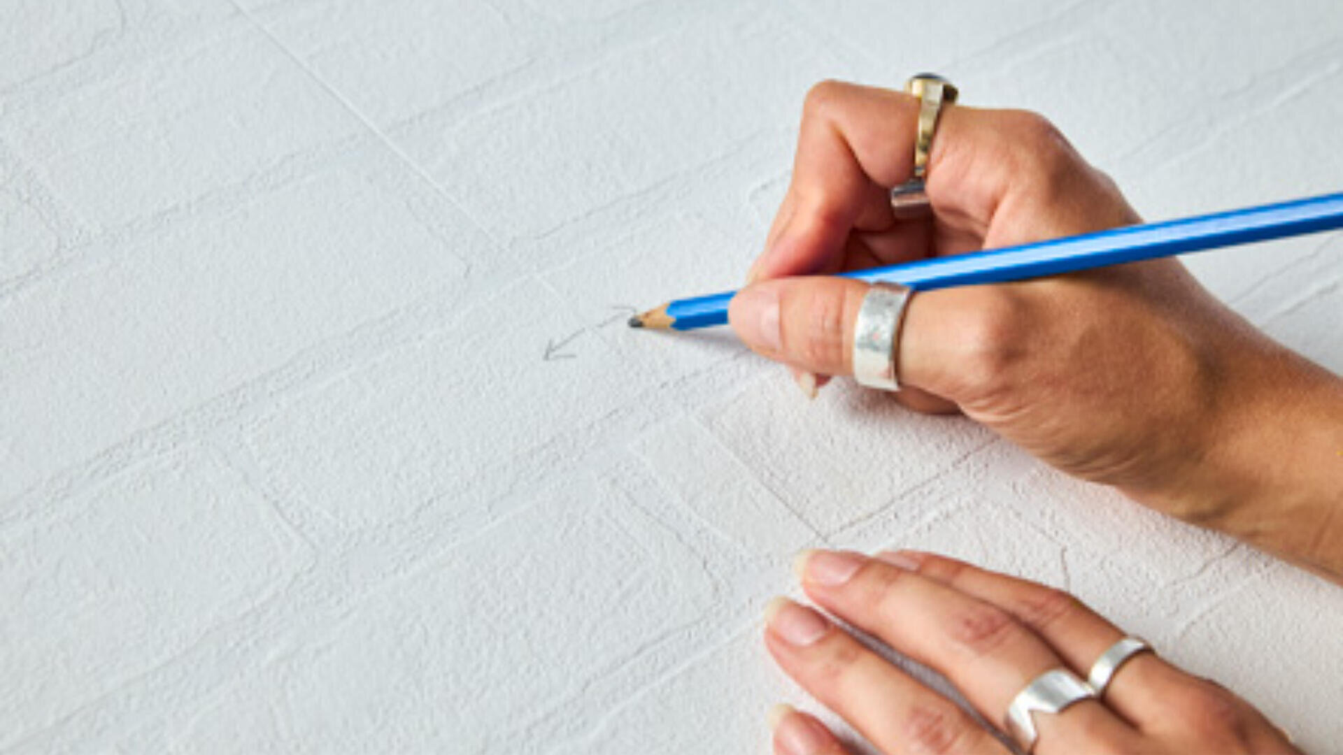 Eine Person mit silbernen Ringen zeichnet mit einem blauen Stift einen Pfeil auf eine weiße, strukturierte Oberfläche, die einer Ziegelwand ähnelt.
