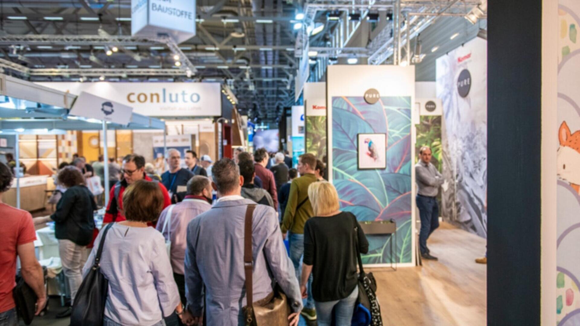 A busy exhibition hall with many people walking and interacting, surrounded by booths displaying signs, posters, and products under bright overhead lighting.