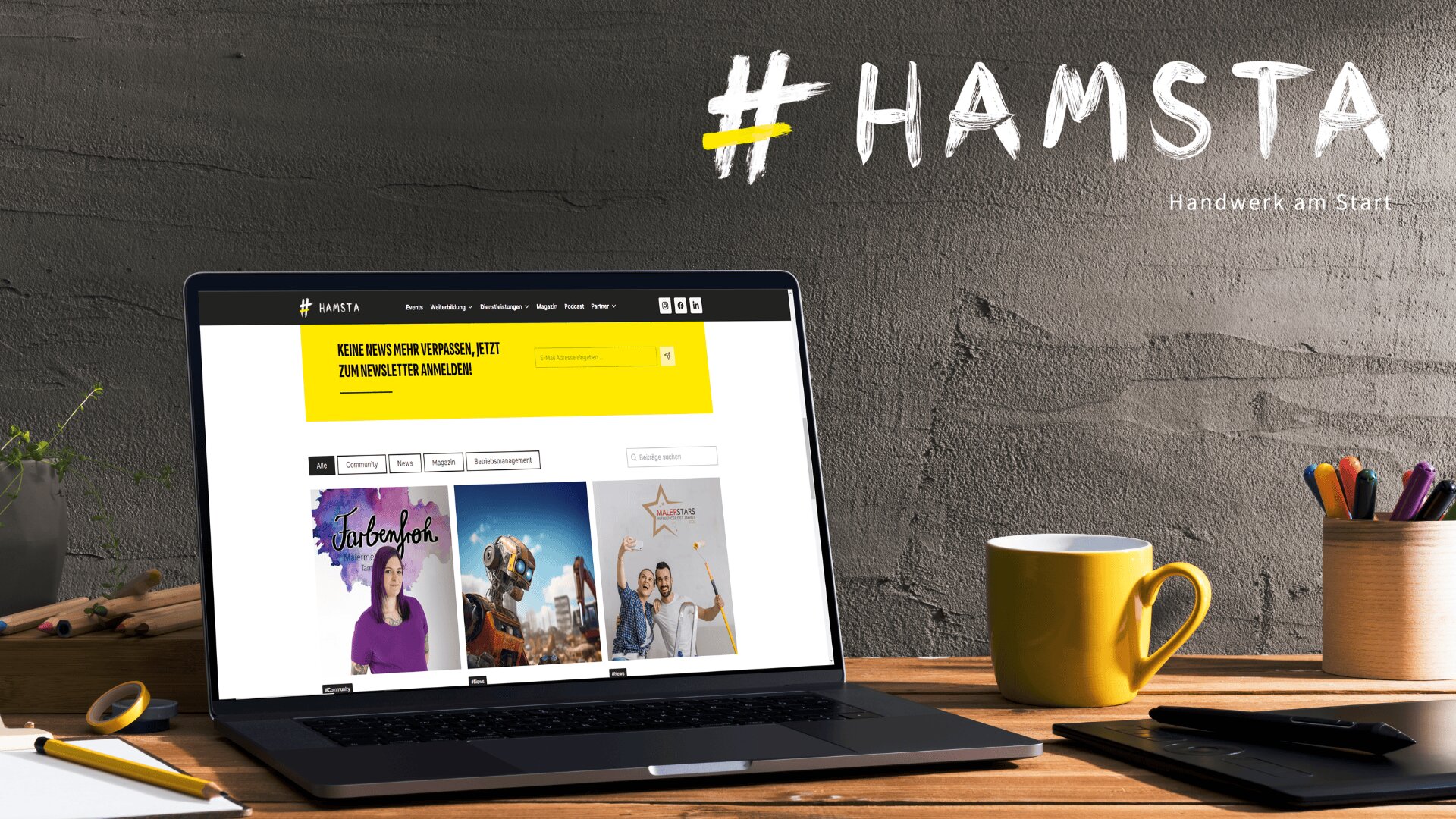 Ein Arbeitsplatz mit einem Laptop, auf dem die HAMSTA-Website angezeigt wird, einer gelben Tasse, Stiften in einem Halter und Pflanzen auf einem Holzschreibtisch. An der Wand steht in großen weißen Buchstaben „#HAMSTA Handwerk am Start.“.