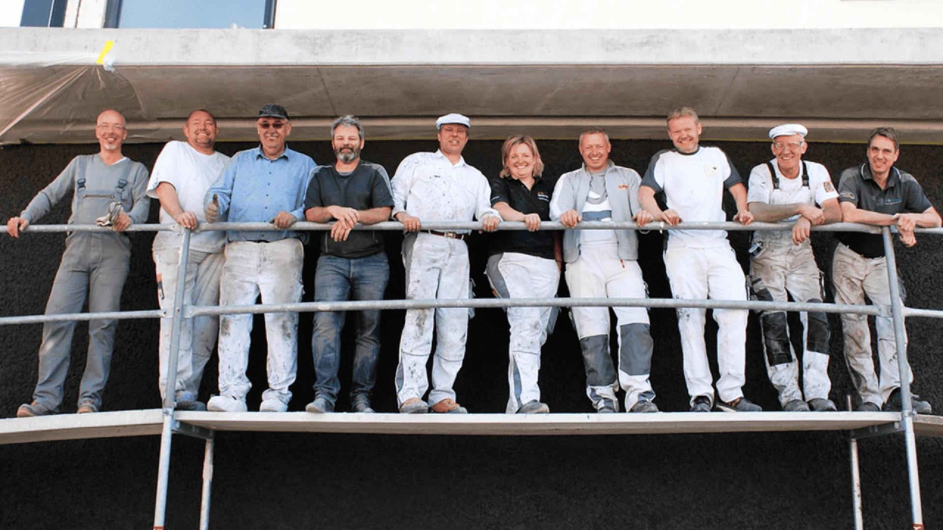 A group of ten people, some in white work overalls and some in casual clothing, stand smiling on scaffolding in front of a building.