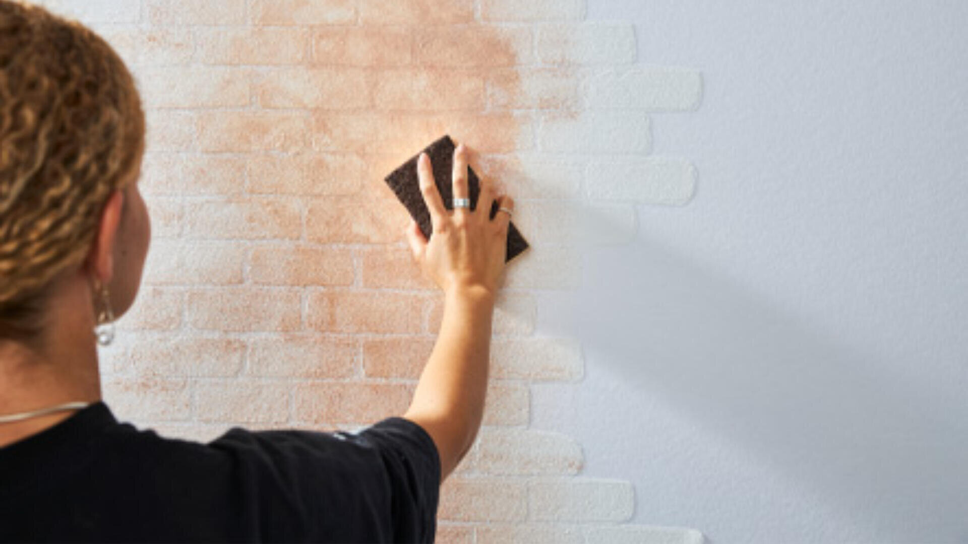 Eine Person trägt mit einem Schwamm eine rotbraune Farbe auf eine weiße Wand mit Ziegelmuster auf und bedeckt dabei die Ziegel teilweise.