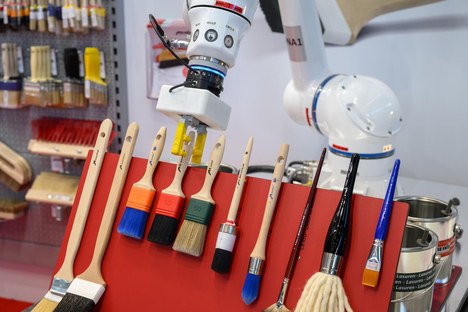 Ein Roboterarm wählt einen Pinsel aus einer Ausstellung verschiedener Pinsel auf einem roten Ständer aus. Im Hintergrund hängen weitere Pinsel an einer Wand.
