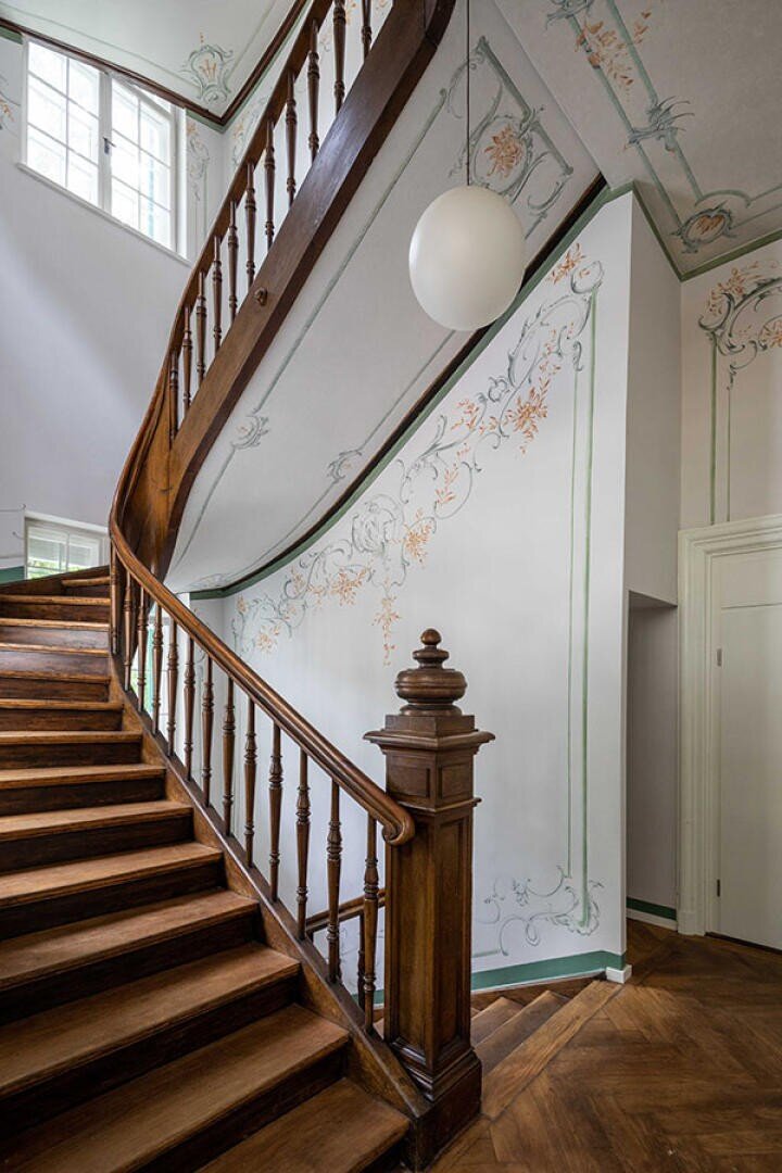 Eine Holztreppe mit verziertem Geländer führt in einem hellen Flur nach oben. Dekorative pastellfarbene Blumenmuster zieren die weißen Wände und die Decke, und über ihr hängt eine runde weiße Leuchte.