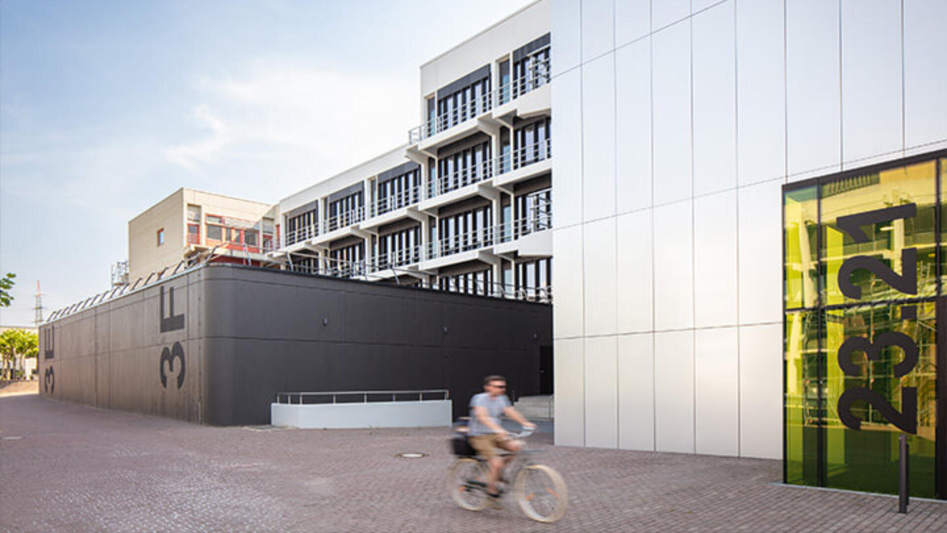 Eine Person fährt mit dem Fahrrad an einem modernen Gebäude mit schwarz-weißer Fassade, großen Fenstern und einem Glasbereich vorbei, der mit den gelben Zahlen 1 bis 4 gekennzeichnet ist. Der Himmel ist klar und sonnig.