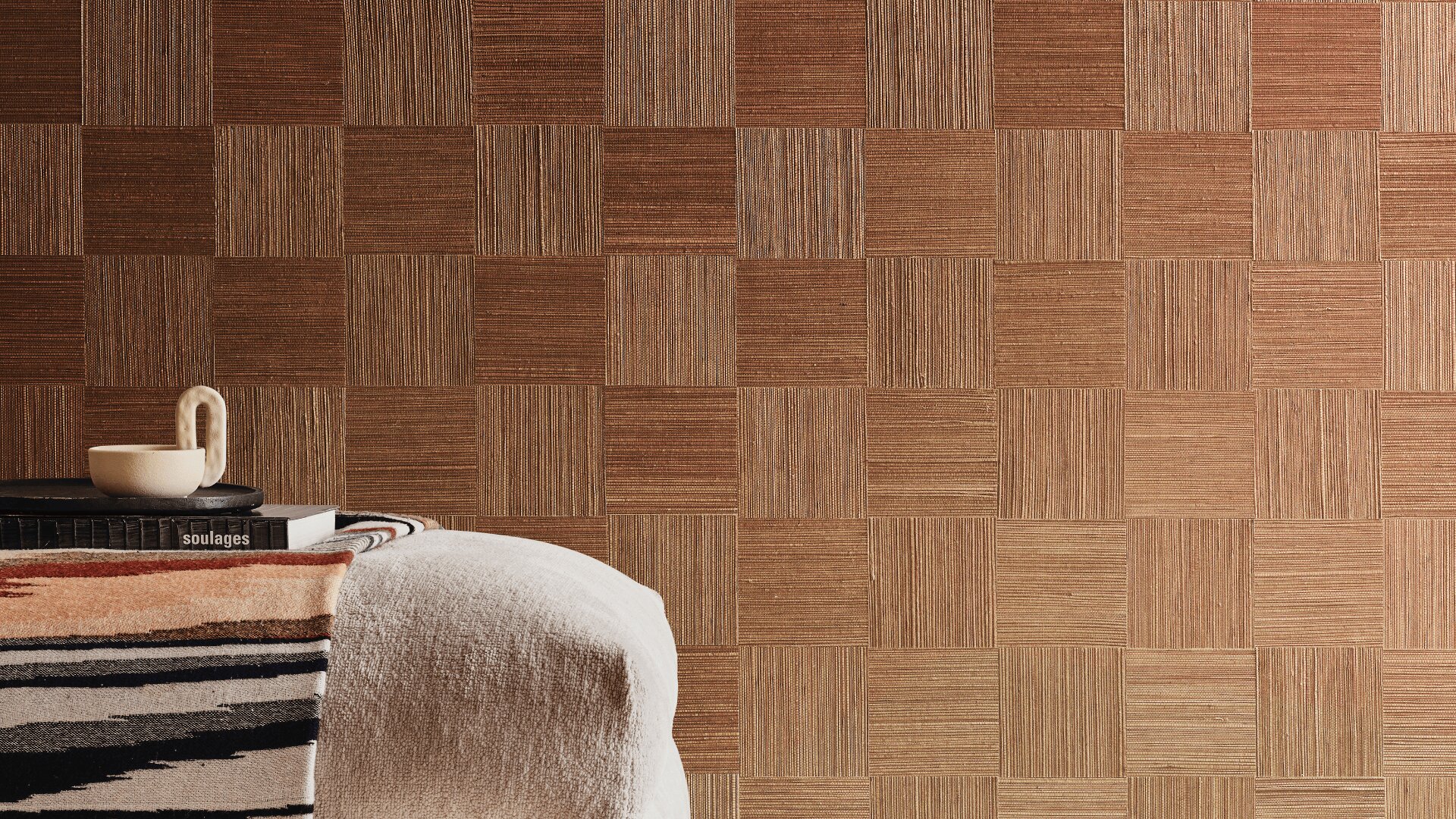 Eine gemütliche Ecke mit einem weißen Sessel mit Strukturmuster, einer gemusterten Decke, einem Stapel Bücher und einer Kaffeetasse vor einer karierten braunen Wand mit Strukturmuster.