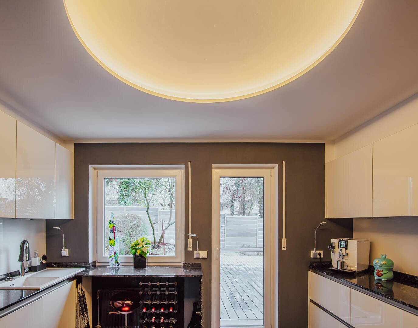 A modern kitchen with sleek white cabinets, black countertops, large windows, a glass door to a deck, and a round, glowing ceiling light. A plant, wine rack, and coffee machine are visible on the counters.