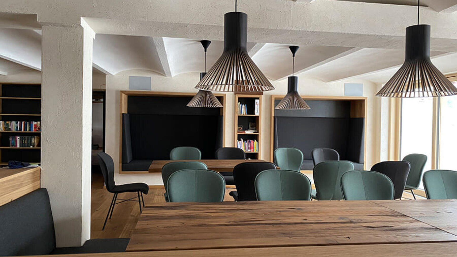 Ein moderner, gemütlicher Besprechungs- oder Arbeitsbereich mit Holztischen, grünen Stühlen, integrierten schwarzen Sitzecken, Bücherregalen und schwarzen Pendelleuchten an der Decke. Natürliches Licht erhellt den Raum.
