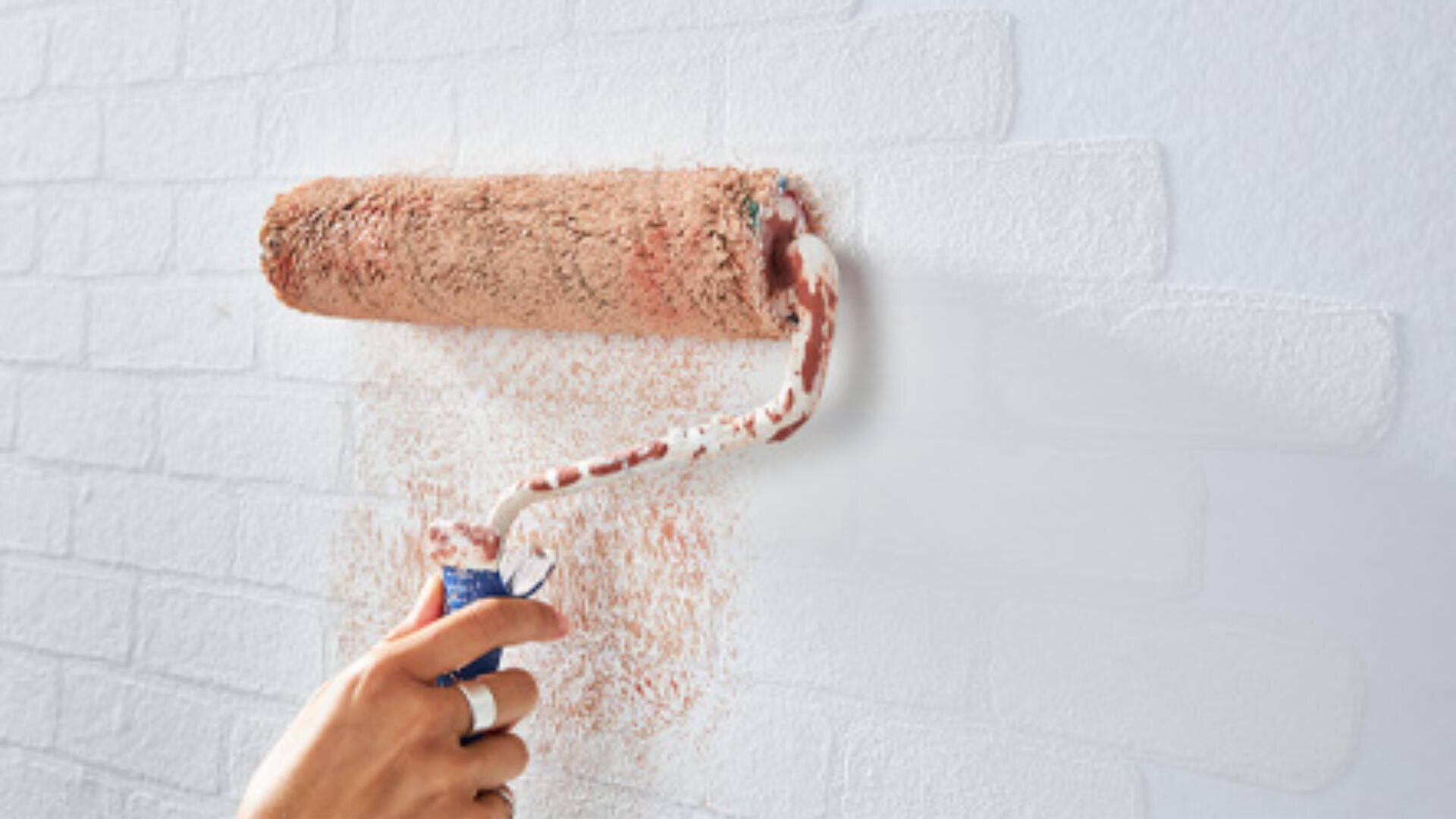 Eine Hand trägt mit einer Farbrolle weiße Farbe auf eine Ziegelwand auf und überdeckt dabei teilweise die ursprüngliche rotbraune Farbe der Ziegel.