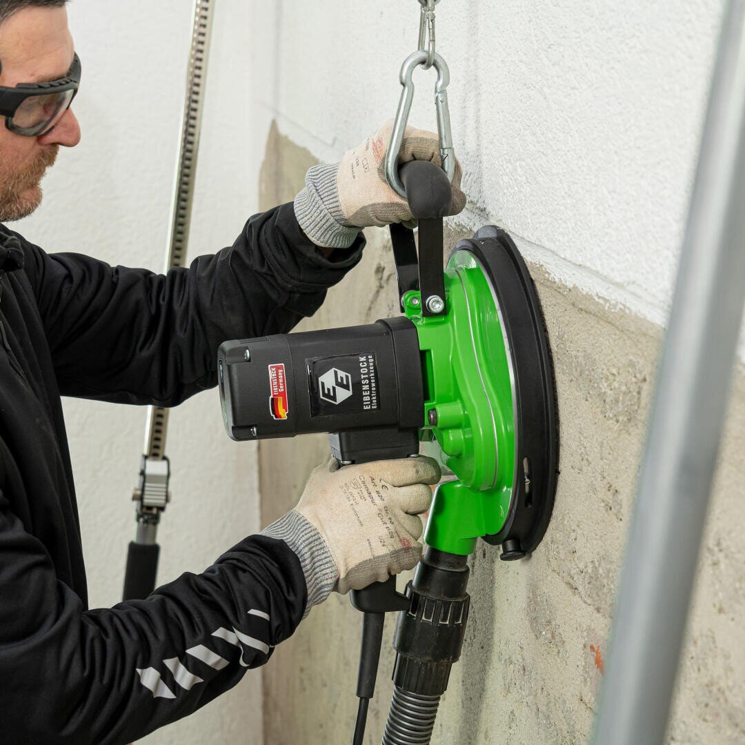 Eine Person mit Schutzbrille und Handschuhen glättet oder schleift mit einem grünen Elektrowerkzeug mit Saugaufsatz eine Betonwand, wobei ein Staubabsaugschlauch an das Werkzeug angeschlossen ist.