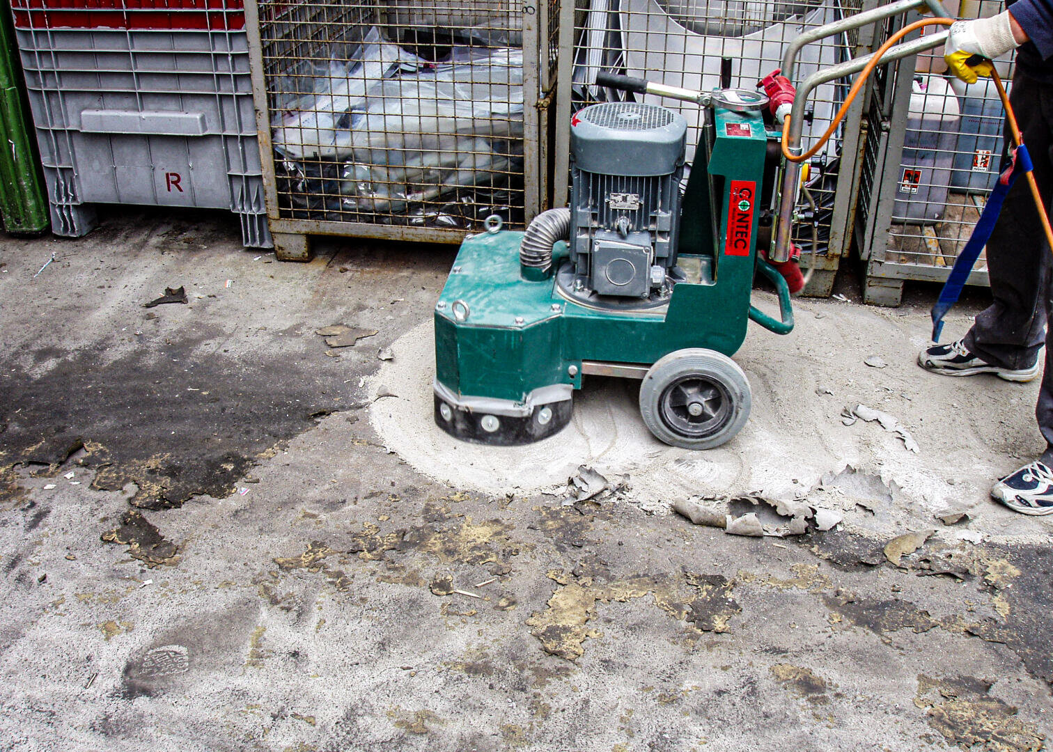 Eine Person entfernt mit einer grünen Bodenschleifmaschine beschädigten Beton von einem rauen Industrieboden. Im Hintergrund sind Schutt und Container zu sehen.