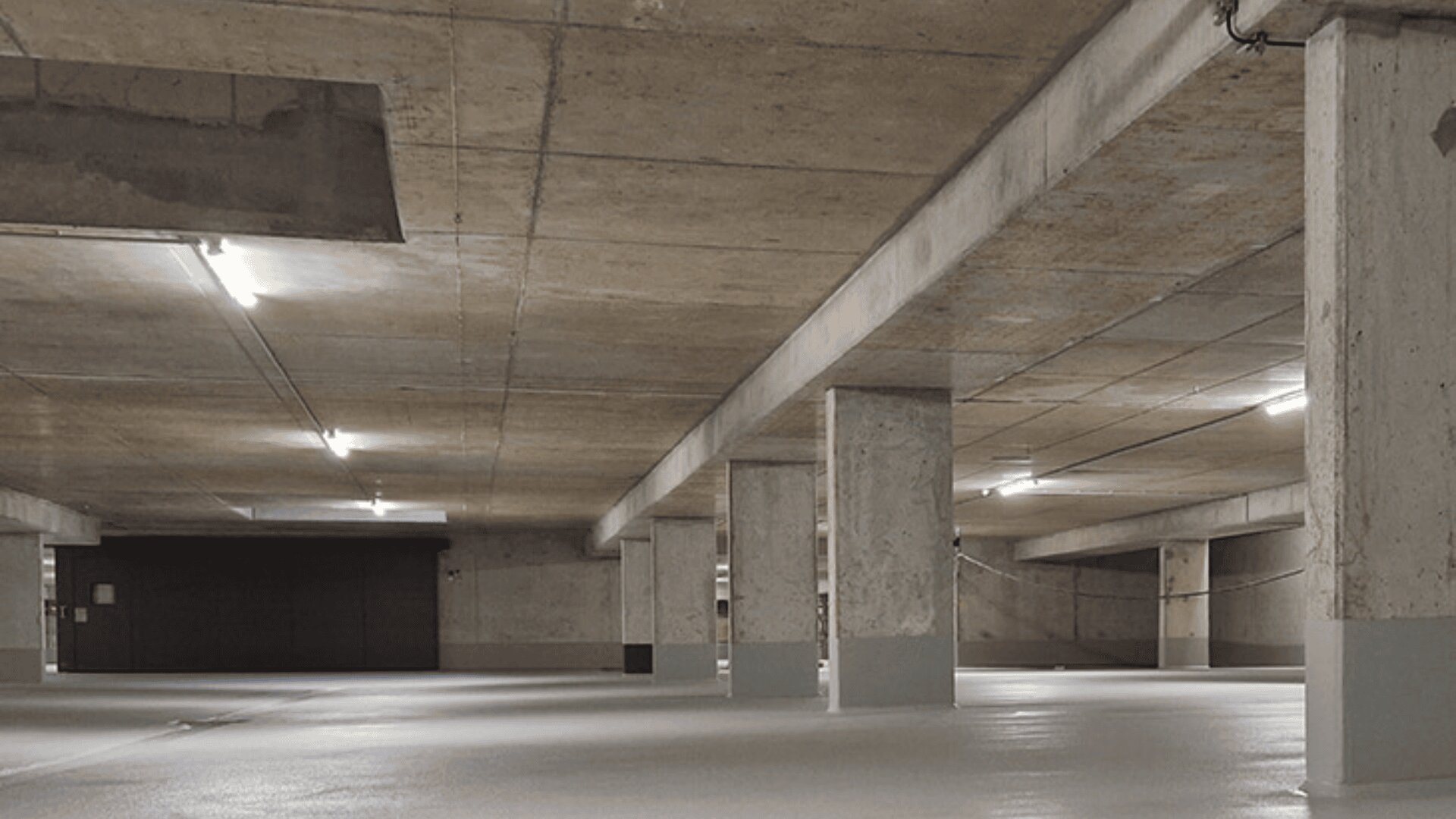 Eine leere Tiefgarage mit Betonpfeilern und -decke, hell erleuchtet von Leuchtstoffröhren. Der Boden ist sauber und glatt, weder Fahrzeuge noch Personen sind zu sehen.