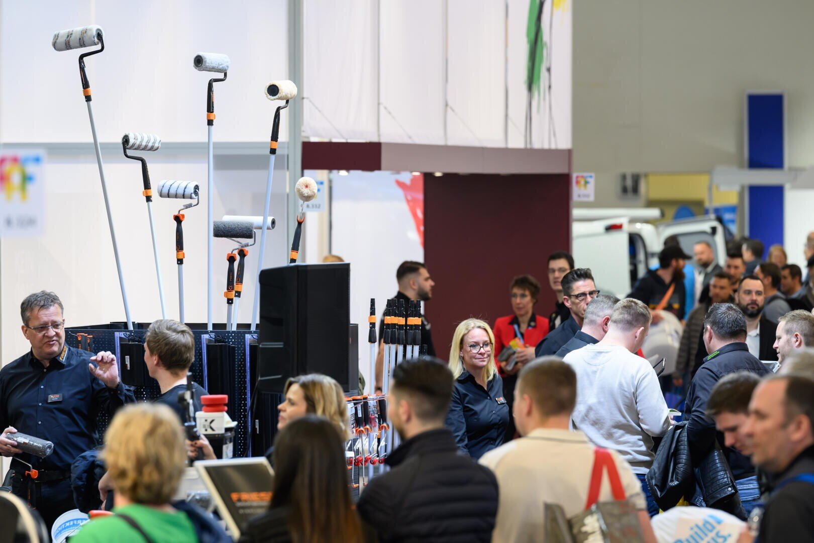 Eine überfüllte Messe- oder Ausstellungshalle mit Menschen, die an Ständen mit Farbrollern und Werkzeugen interagieren. Die Besucher unterhalten sich und betrachten die ausgestellten Produkte.