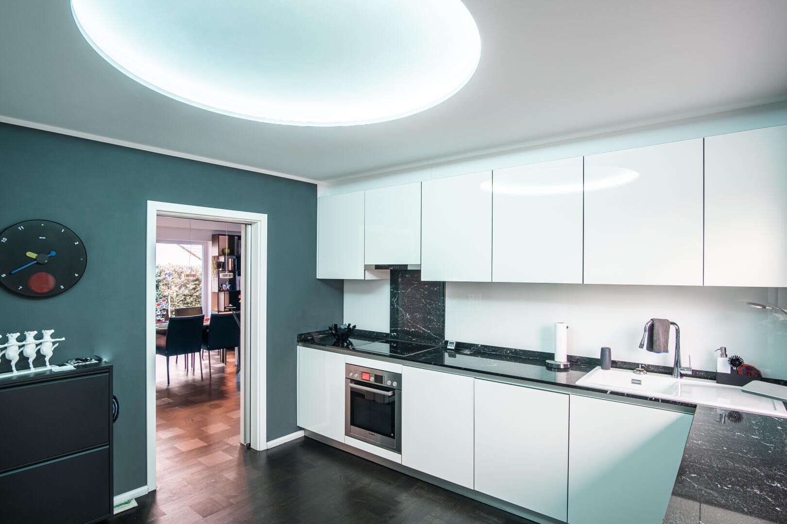 Moderne Küche mit eleganten weißen Schränken, schwarzen Arbeitsplatten, eingebautem Backofen und Wasserkocher. Eine große runde Deckenleuchte erhellt den Raum. Eine offene Tür führt zum Essbereich mit Holzboden.