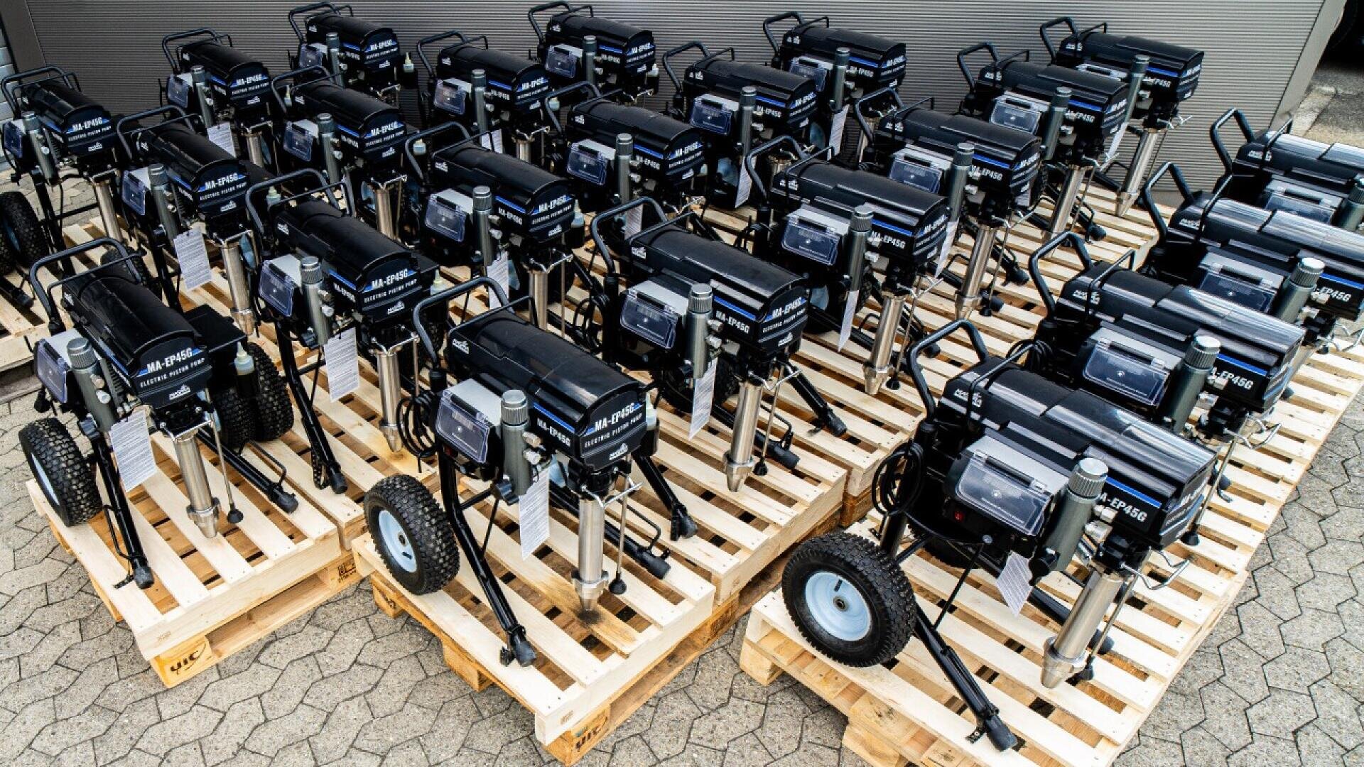 Rows of black industrial machines with wheels are neatly arranged on wooden pallets outdoors, ready for transport or storage. The machines are identical and aligned in a grid pattern on a paved surface.