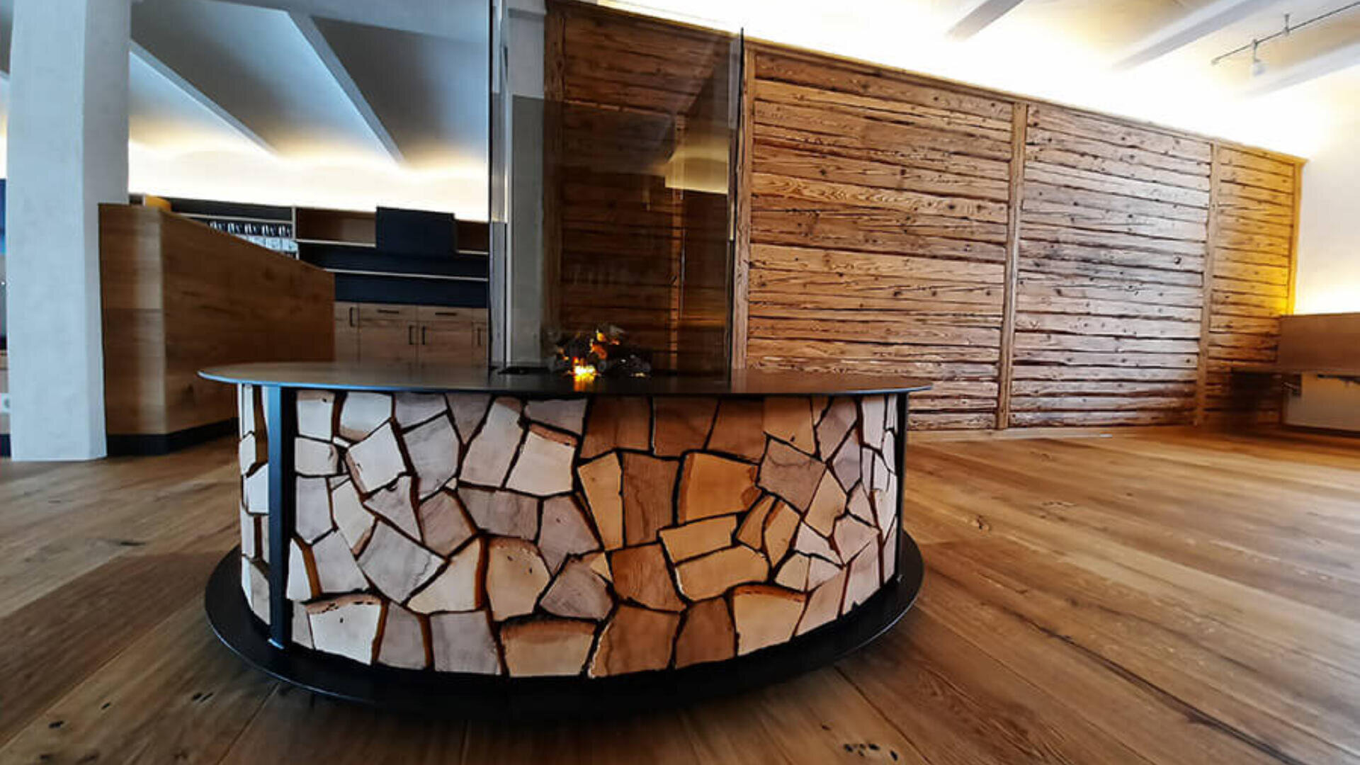 Ein moderner, runder Kamin mit einer Frontplatte aus unregelmäßigen Holzstücken steht in einem großzügigen Raum mit Holzböden und Wänden aus horizontalen Holzbrettern.