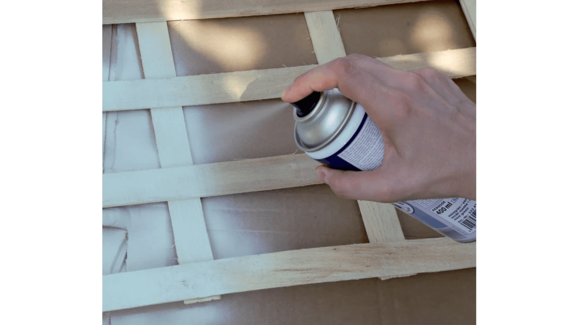Eine Hand hält eine Sprühdose und sprüht weiße Farbe auf ein Holzgitter, das über einer Kartonoberfläche liegt.