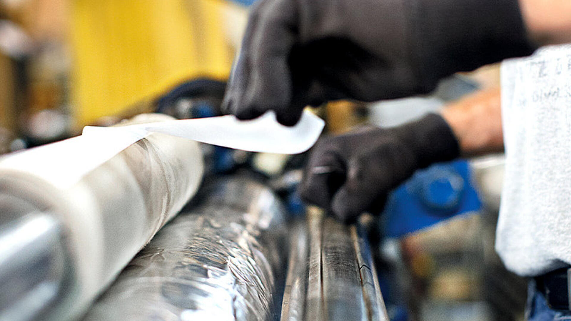 Eine Person mit schwarzen Handschuhen bedient Maschinen und bringt in einer industriellen Umgebung eine von einer großen Rolle abgewickelte Plastikfolie an oder prüft sie.