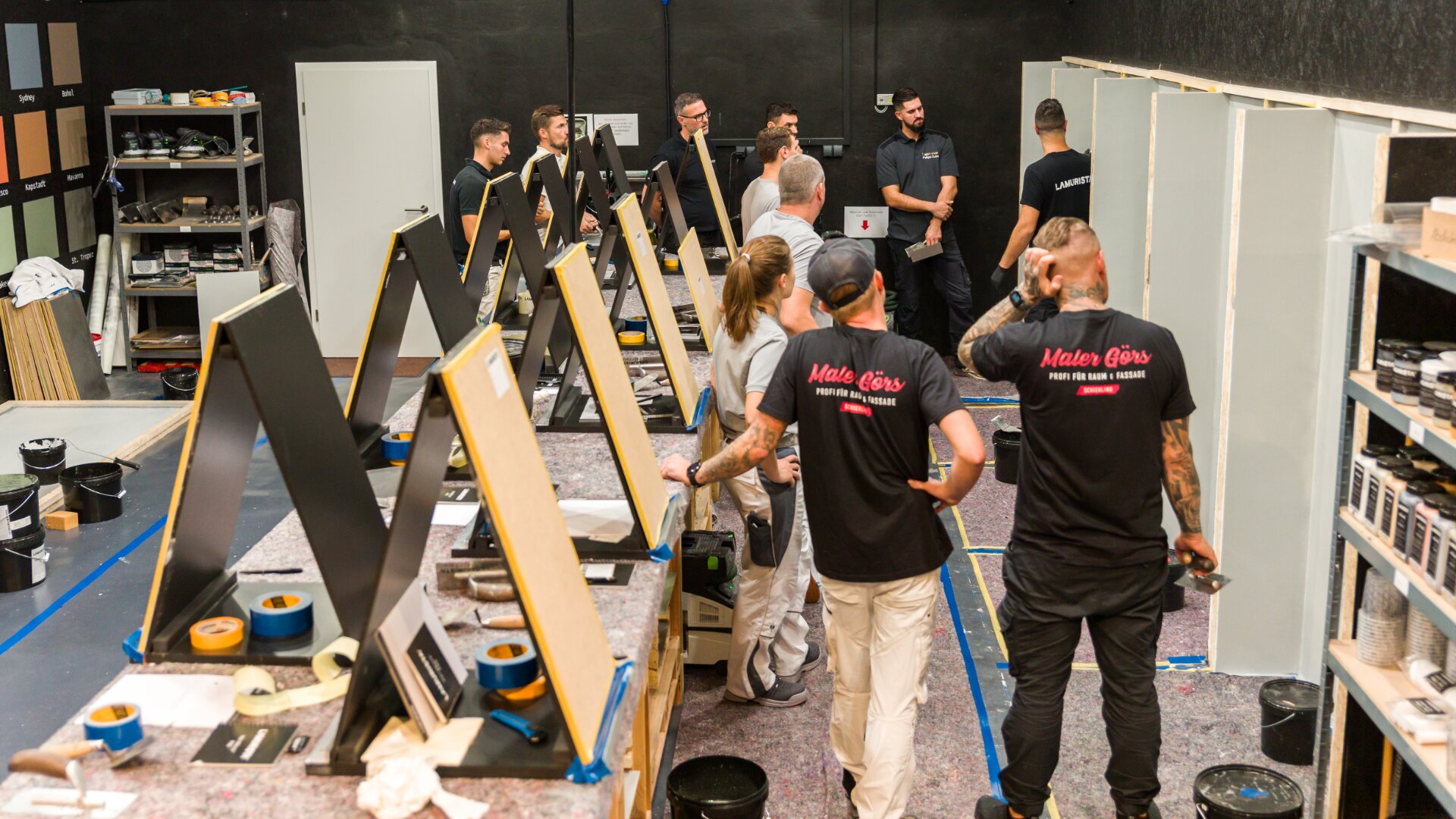 Eine Gruppe von Menschen arbeitet und diskutiert in einem Werkstattraum. Auf den Tischen sind bemalte Tafeln aufgestellt, überall liegen Malutensilien herum und mehrere Menschen tragen passende schwarze Maler-Gåris-Shirts.