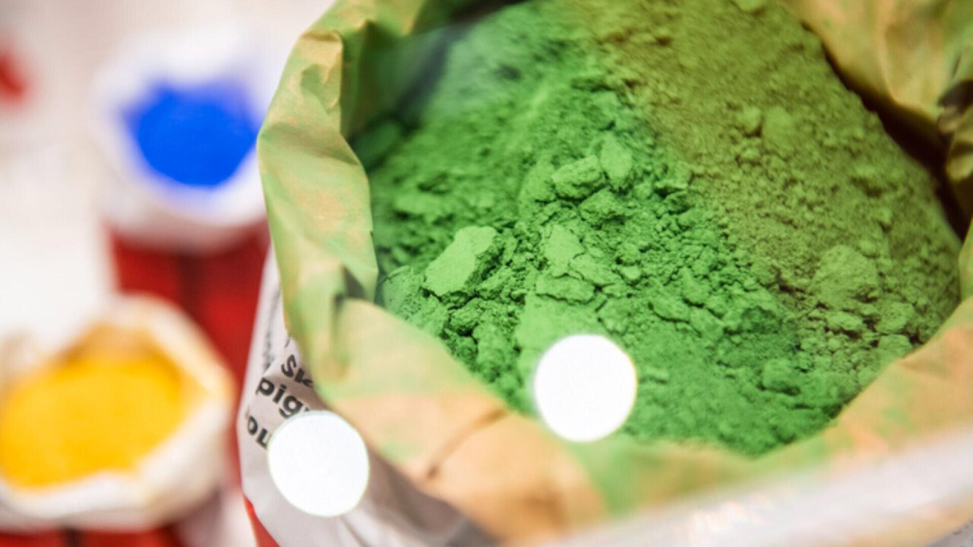 A close-up of a paper bag filled with bright green powder, with blurred blue and yellow powders in sacks visible in the background.