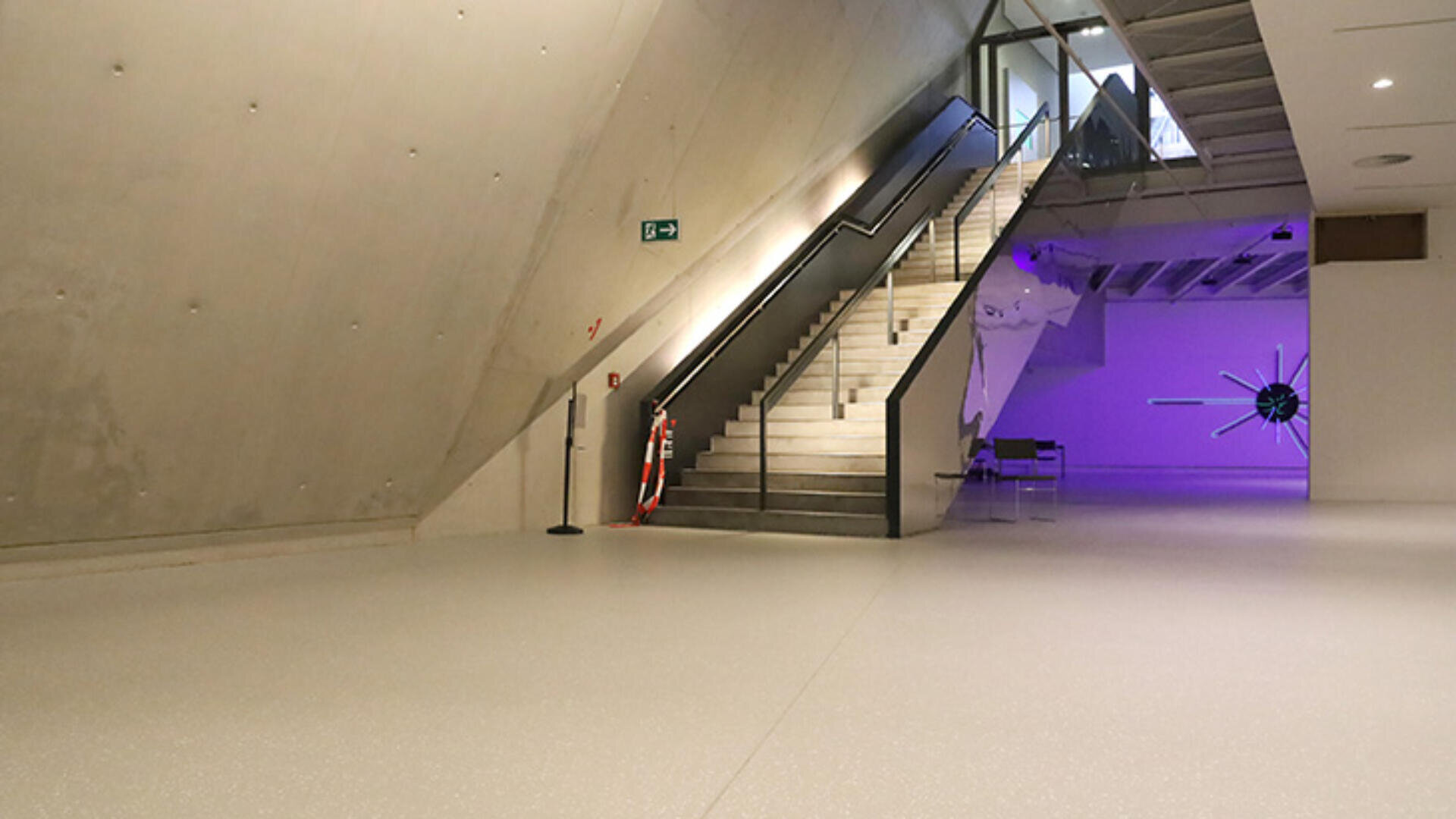 Ein modernes, minimalistisches Interieur mit Betonwänden, einer breiten Treppe, die in eine obere Etage führt, und violetter Beleuchtung im Hintergrund, die eine große, abstrakte Skulptur hervorhebt.